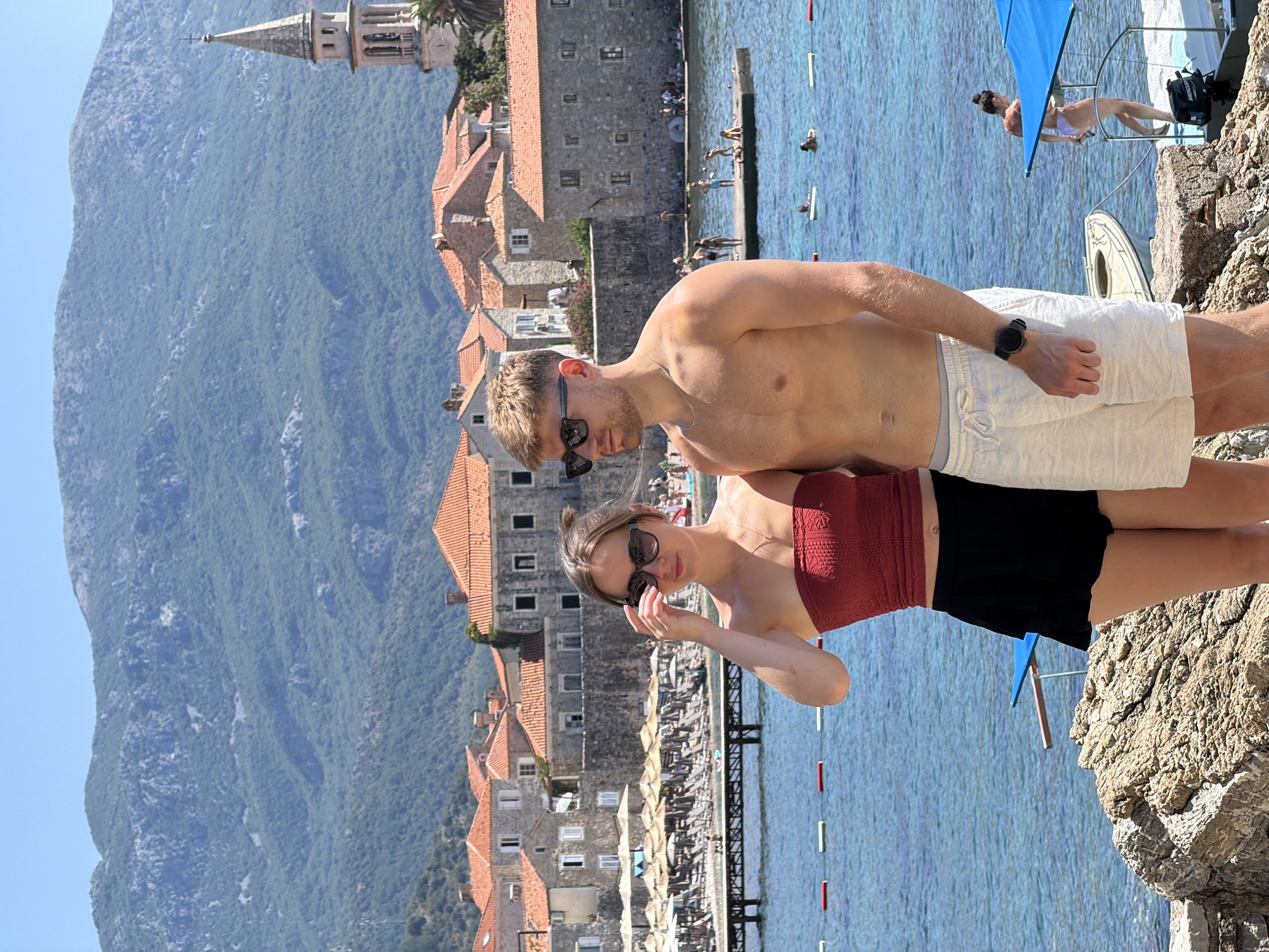 In Budva, Montenegro, a young couple poses for a photo in front of a coastal town scene. The man, wearing white shorts and no shirt, stands on the right, with his left arm slightly bent. The woman, on the left, wears a red bandeau top and black shorts, and adjusts her sunglasses with her right hand. They are positioned on a rocky shore overlooking the clear blue Adriatic Sea. In the background, the town of Budva features characteristic red-tiled roofs, stone buildings, and a prominent bell tower, with lush green mountains rising behind it. Beach umbrellas and loungers are visible further down the coastline, and several people are scattered in the water and on a small pier. The scene is bathed in bright sunlight, suggesting a warm, summer day.