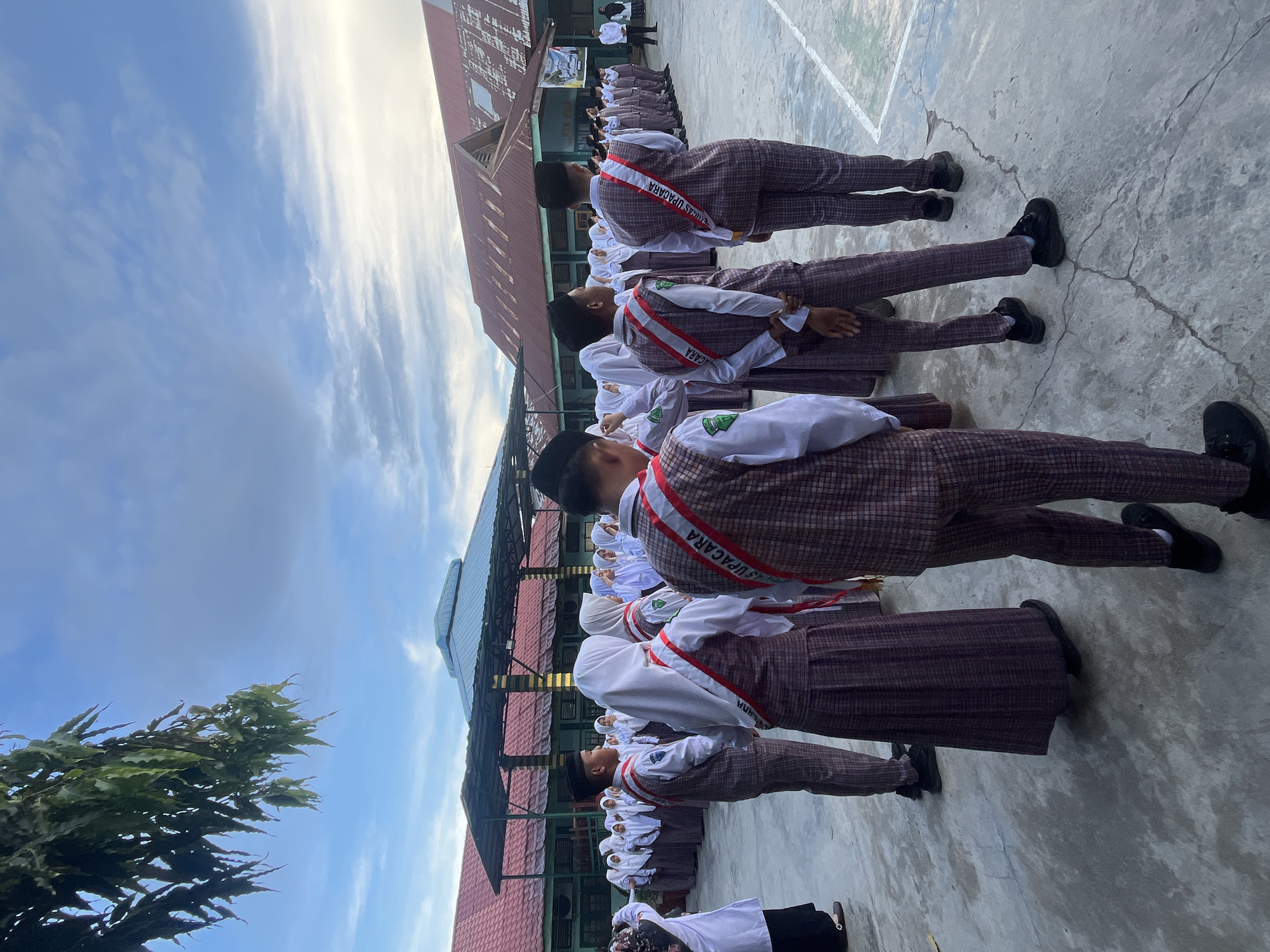 The image shows a group of uniformed students standing in a schoolyard in Lhokseumawe, Indonesia. The students are wearing white shirts, plaid vests and pants or skirts, and some are wearing black caps. They are also wearing red and white sashes across their chests, some of which have text visible. In the background, there are school buildings with red roofs and awnings. The sky is blue with some clouds, and a tree branch with green leaves hangs from the top left corner. The students appear to be in formation, possibly for an assembly or event.