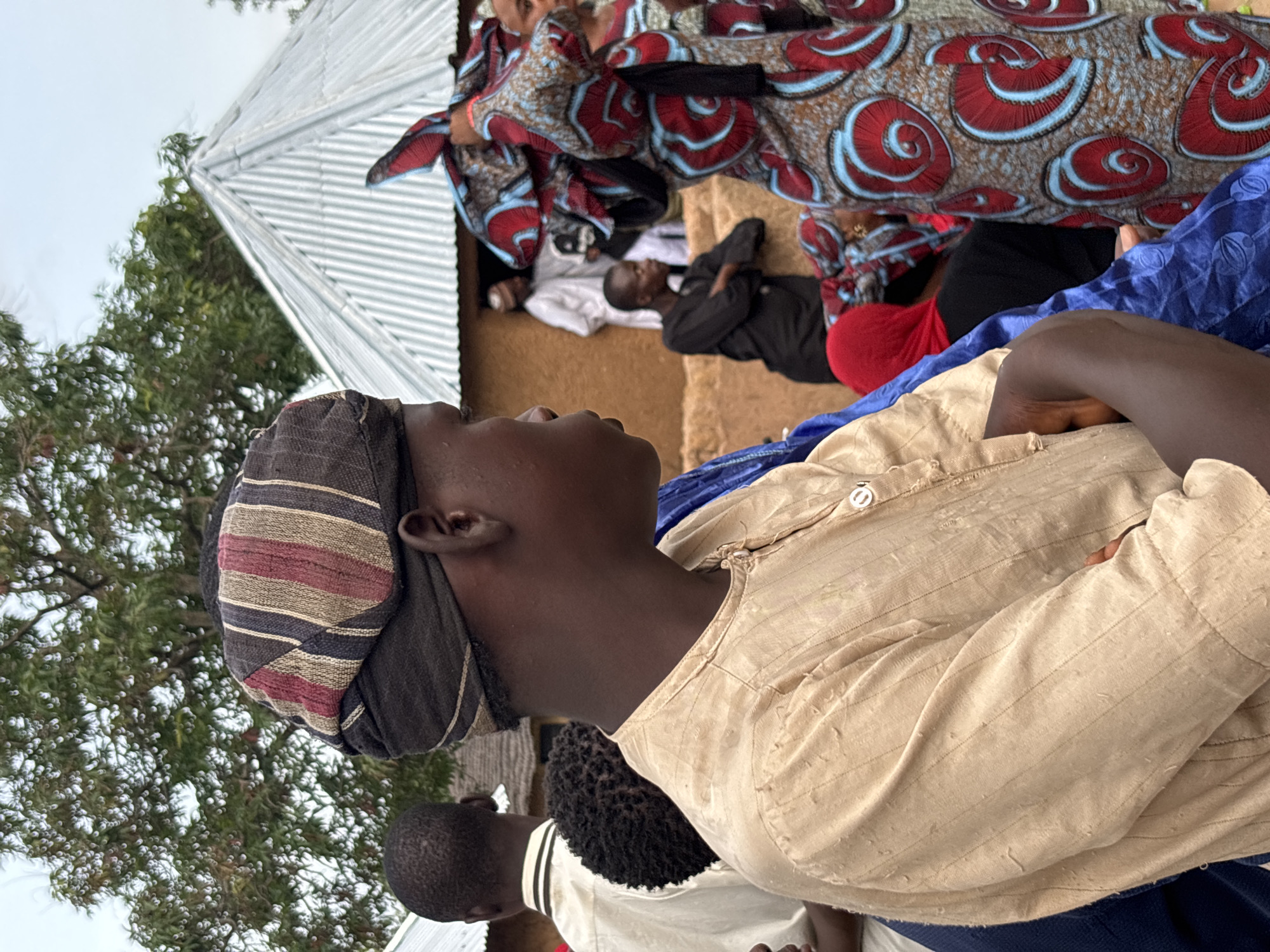 In Jos, Nigeria, a medium shot captures a gathering of people outdoors. In the foreground, a young man with dark skin is shown from the chest up, facing to the right. He wears a traditional tan garment with vertical, subtle stripes and a dark head wrap with colorful bands of red, brown, and black. His arms are crossed in front of him. Behind him, other individuals are visible. To his left, a man in a white shirt is partially visible. To his right, a woman is adorned in a vibrant, patterned fabric featuring large red and blue swirls. In the background, traditional mud-brick buildings and a corrugated metal roof are apparent. A large tree with green foliage looms over the scene. The overall atmosphere suggests a community event or gathering.