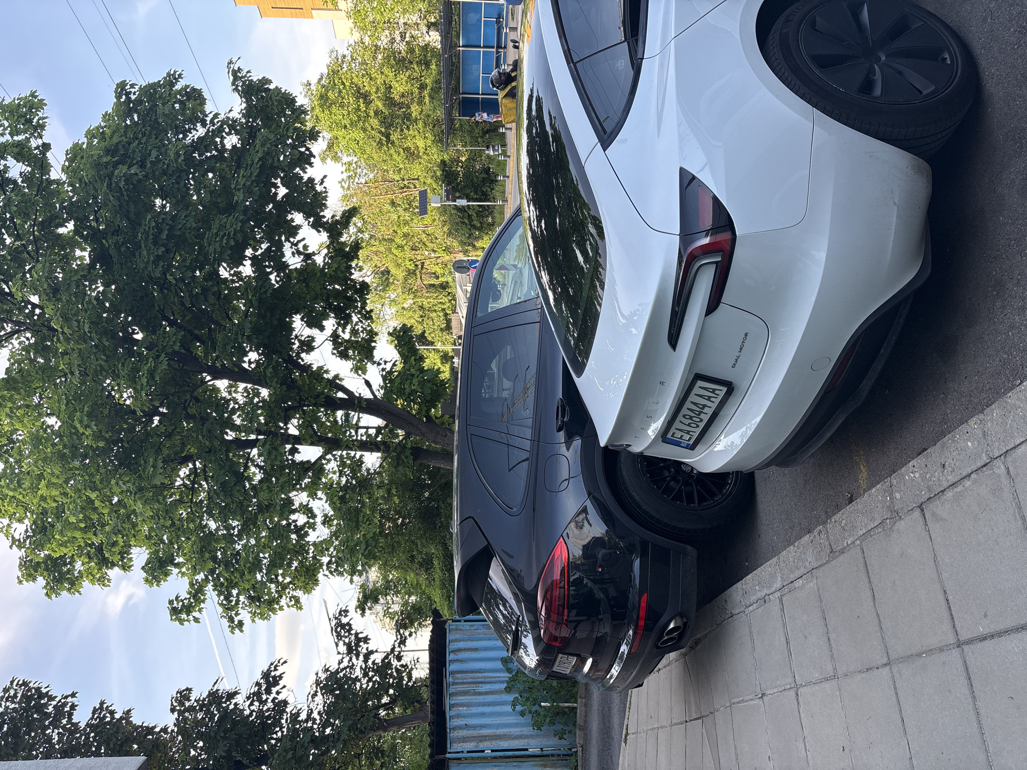 The image depicts two cars parked on a city street in Sofia, Bulgaria. The first car is a dark-colored Porsche Cayenne SUV. The second car is a white Tesla Model Y. The Tesla's license plate is EA 6844 AA. Both cars are parked near a sidewalk and curb. In the background, a large tree, building, bus stop, and street sign can be seen. The sky is visible in the upper left corner and appears partly cloudy. A blue corrugated metal structure is seen at the bottom of the image, left of the Porsche. The photograph appears to have been taken during the daytime.