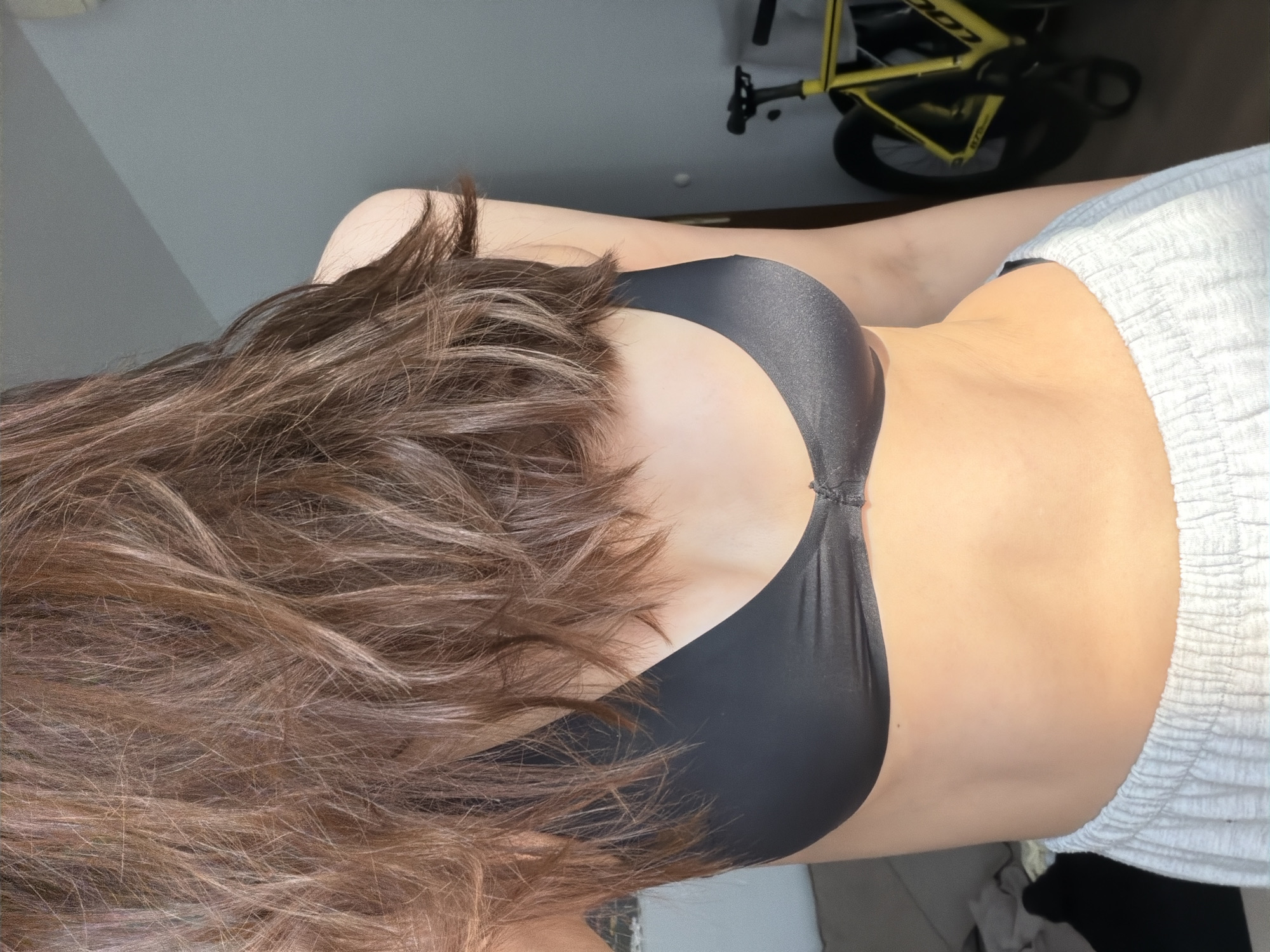 A person wearing a black sports bra and light grey sweatpants stands in a room, facing to the left of the frame. Their mid-torso and long, brown, wavy hair are visible. Sunlight streams in from the right, illuminating the person's skin and creating a soft glow. In the background, a bright yellow bicycle with thick tires is leaning against a grey wall. The environment appears to be residential, possibly an apartment in Khimki, Russia, given the natural light and indoor setting.