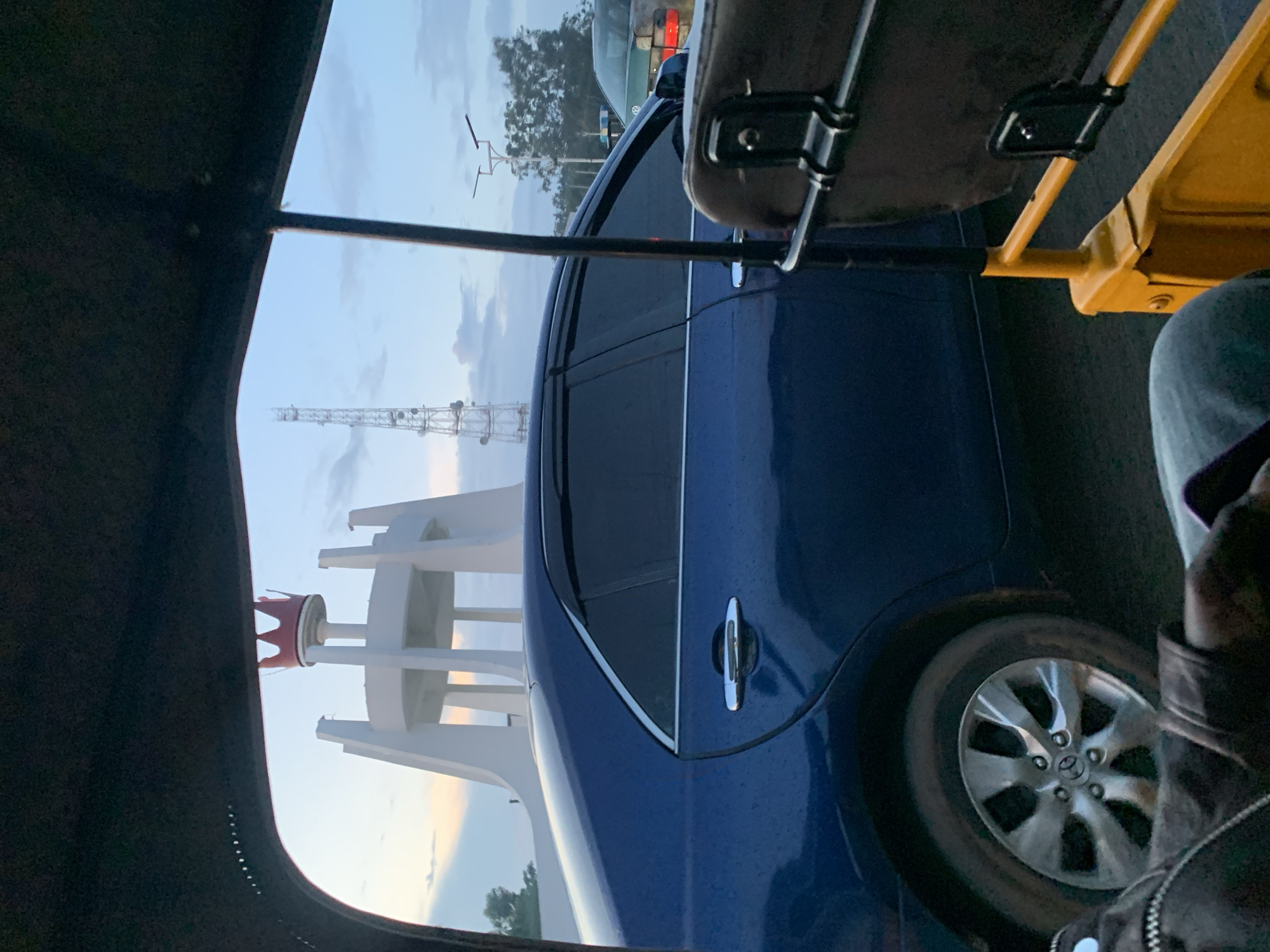 The image captures a view from inside a yellow auto-rickshaw, looking outwards towards a cityscape in Jos, Nigeria. The prominent structures visible are a large white tower with a red crown-like top, a tall telecommunications tower, and several vehicles, including a dark blue sedan and a green car. The sky is overcast with clouds, suggesting it is either early morning or late evening. The blue sedan is parked or moving slowly on a paved road, and its reflections on the wet surface indicate recent rain. The auto-rickshaw's yellow frame and black seating are partially visible in the foreground. The overall scene conveys a sense of urban life in Jos, with elements of transportation, architecture, and infrastructure.