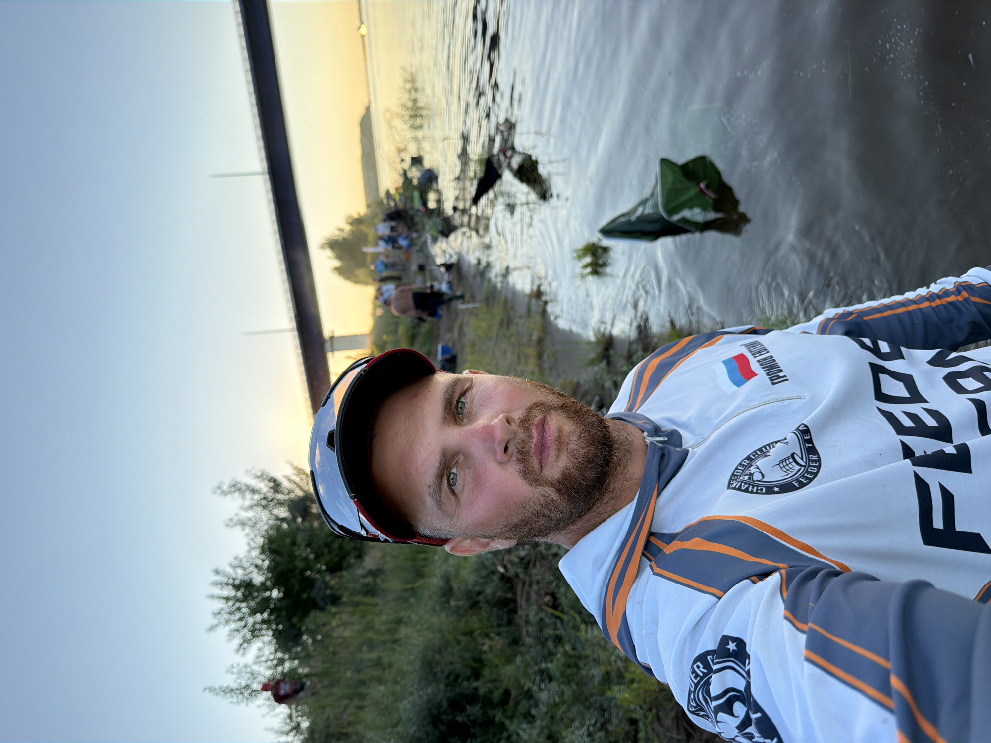 A man is taking a selfie by the river in Tarasovo, Russia. He is wearing a white and gray long-sleeved shirt with orange accents and has a beard and a baseball cap with sunglasses on top. The river is calm and reflects the clear sky. In the background, there are several people on the riverbank, some of whom appear to be fishing. A bridge spans the river in the distance. The sun is setting, casting a warm glow over the scene. There is a green fishing net in the water near the man.