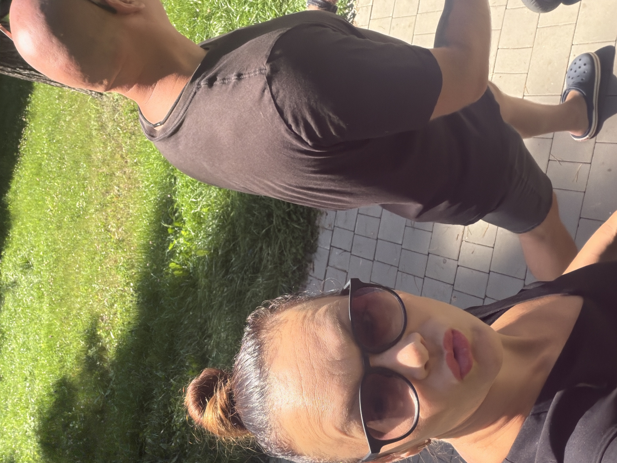 In Votkinsk, Russia, on a sunny day, a woman with her hair in a bun and wearing sunglasses and a black v-neck shirt is looking at the camera with her lips pursed. Beside her and to her right, a bald man wearing a dark t-shirt and dark shorts is walking. They are on a paved walkway, with green grass and a tree to their left.