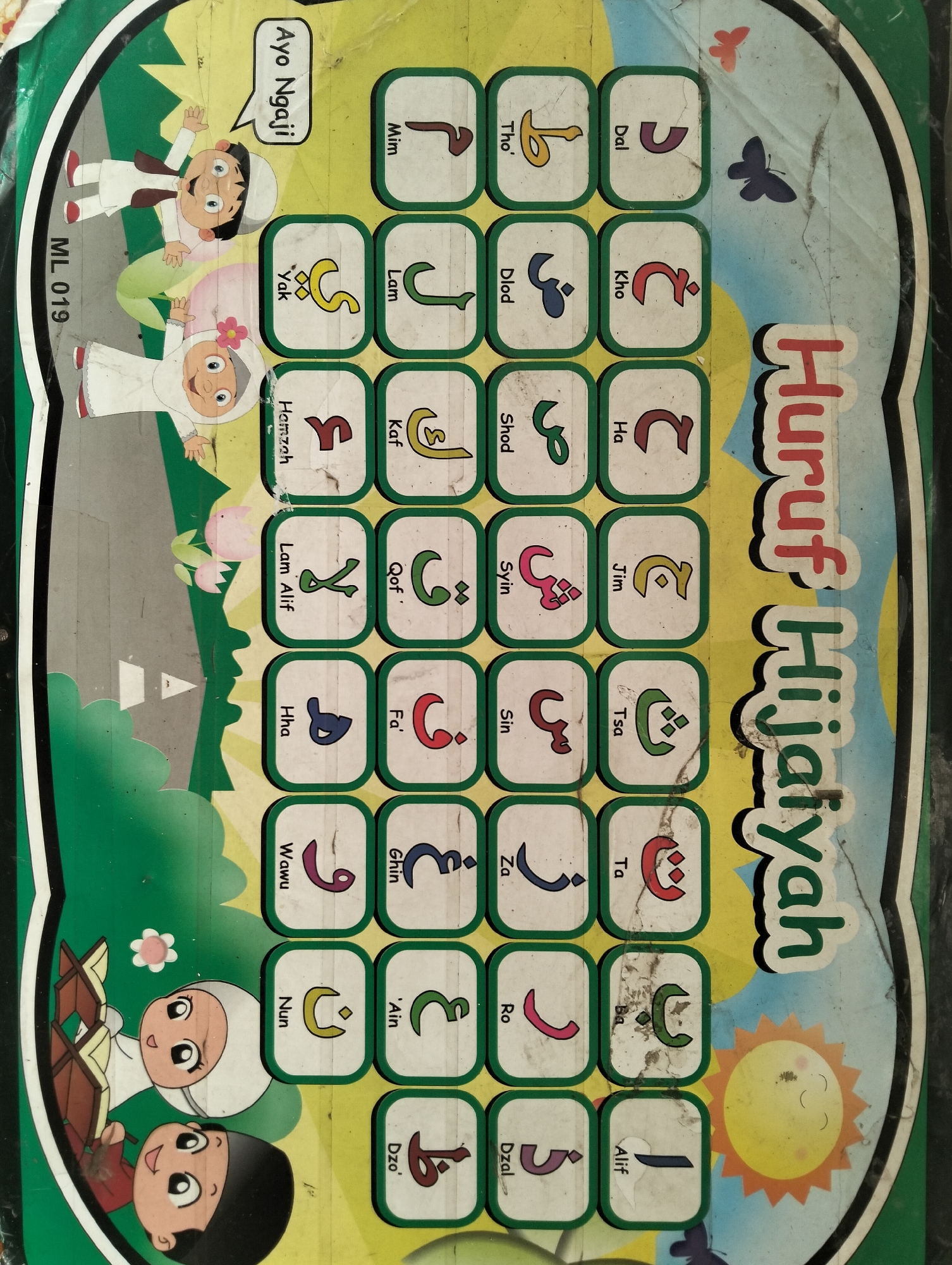 The image is a vertically oriented photograph of a children's educational toy, specifically an Islamic alphabet learning board. The board features cartoon illustrations of two children, a boy and a girl, dressed in traditional Islamic attire, appearing to be engaged in learning. The background depicts a green landscape with a hint of a road. The main focus is a grid of square buttons, each displaying an Arabic letter with its romanized name above it. These buttons are arranged in columns and rows. On the right side of the board, large, colorful text reads "Huruf Hijaiyah," which translates to "Arabic Letters." There are also decorative elements such as butterflies and a sun. The toy appears to be made of durable material suitable for children. The image has a slightly worn and faded quality, suggesting it has been used. The location provided, City of Medan, Indonesia, is contextual information that cannot be visually verified from the image itself.
