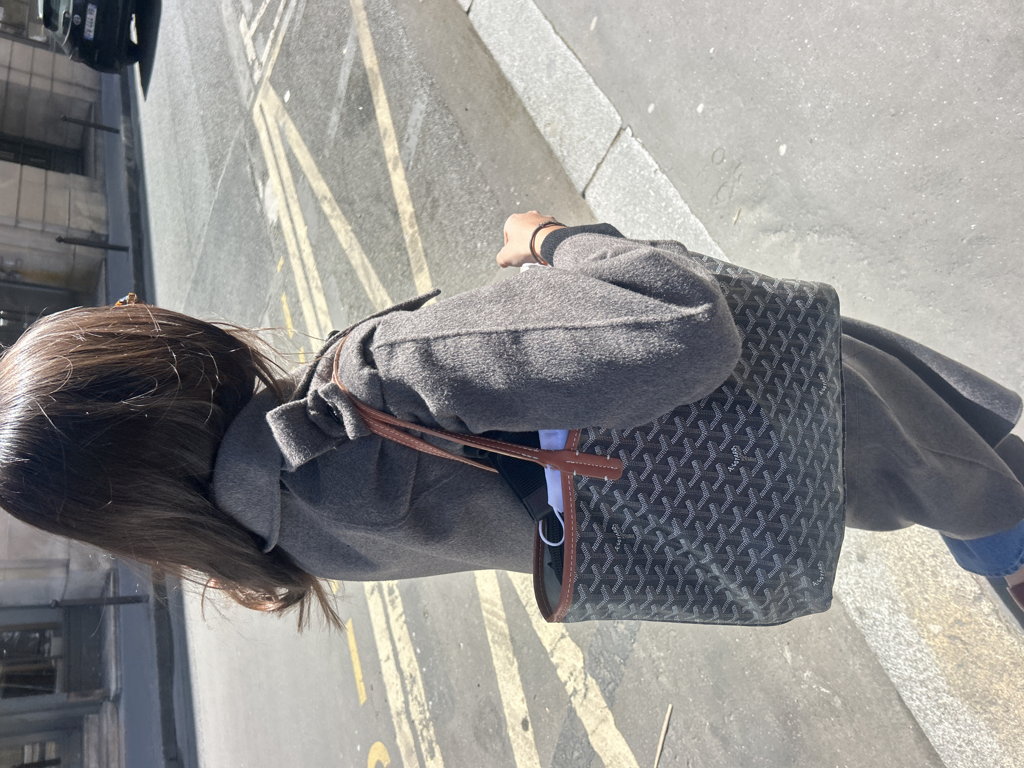 A woman wearing a gray coat and carrying a Goyard handbag is walking on a street in Unknown, Unknown. The street has yellow lines painted on the pavement. A car is parked on the side of the street. The environment appears to be urban.