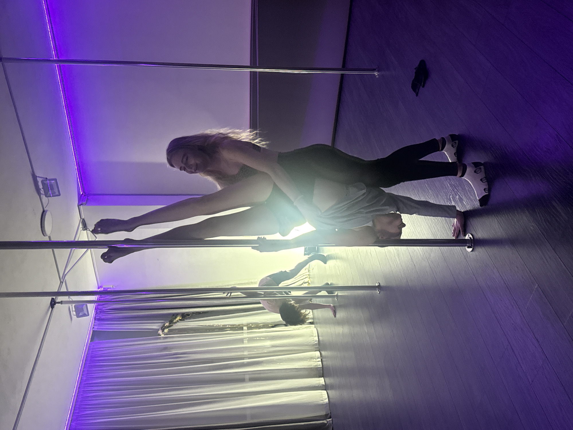 This image captures a dynamic scene inside a pole fitness studio in Batumi, Georgia, characterized by vibrant purple lighting. Two women are prominently featured, engaged in a partner or duo pole dancing move.
The woman on top has long blonde hair and is wearing a patterned top and dark leggings. She is captured mid-pose, extending one leg high up a chrome pole while her other leg wraps around the torso of the woman below. She is smiling, indicating focus and enjoyment. The second woman, with darker hair, is positioned on her back on the dark floor, supporting the woman above her. She wears a light-colored top, dark leggings, and white socks with patterns. Her face is partially visible, showing concentration.
In the background, another pole is visible, and a third person, possibly a child or a smaller adult, is seen practicing an inverted pose on it, with their head facing downwards. The studio floor is dark, possibly made of wood or laminate, reflecting the purple light from the LED strip lights that run along the ceiling. A long white curtain or drape is also visible in the background, near which a small light fixture is mounted on the wall. A small black object, possibly an item of clothing or a shoe, rests on the floor in the upper right portion of the frame. The overall atmosphere is energetic and focused, highlighting the athleticism and artistry of pole fitness. There is no visible text in the image.