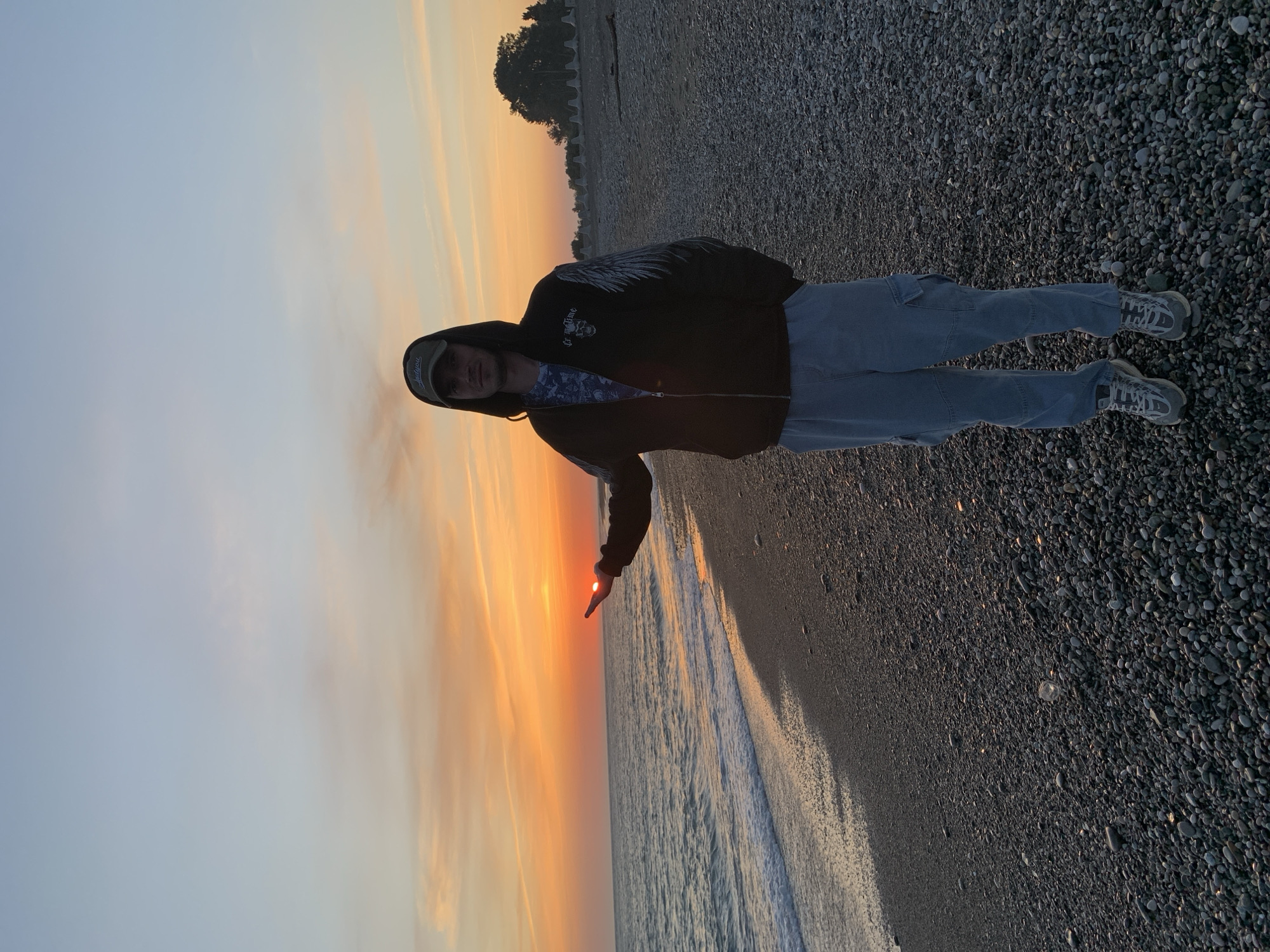 A person wearing a hooded jacket, light-colored pants, and grey sneakers stands on a pebbled beach in Inkiti, Abkhazia. The person is posed so that their index finger appears to touch the setting sun. The background consists of the sunset over the ocean, the shoreline, and a small amount of greenery in the distance.