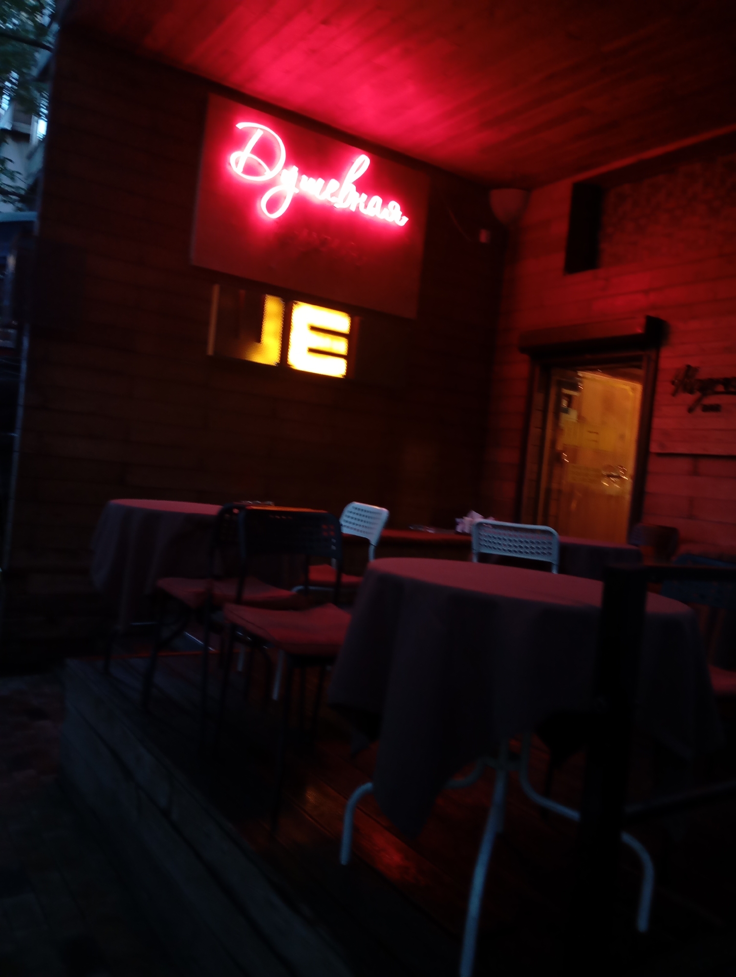 The image shows an outdoor seating area of a establishment at night, illuminated by red neon lighting and a yellow illuminated sign. In the foreground, there are several tables with dark red tablecloths and chairs, some of which are black and others are white. A door with a glass panel and an arched top is visible in the background, with light emanating from within. A neon sign with Cyrillic script, appearing to read "Душевная," is prominently displayed on the wooden wall. To the right of the door, a sign with ornate lettering is mounted on the wall. The overall ambiance is that of a dimly lit, somewhat moody, outdoor bar or cafe. The specific location in Almaty, Kazakhstan cannot be determined from the image alone.