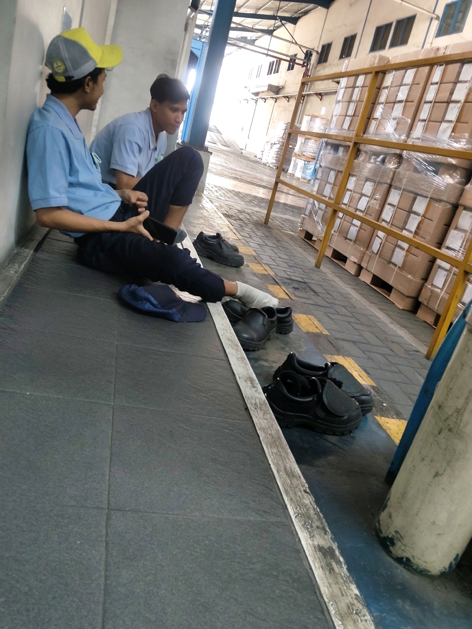 Two young men, dressed in blue shirts and dark pants, are resting in an industrial setting. One man, wearing a yellow and grey baseball cap, is leaning against a light-colored wall and holding a dark rectangular object, possibly a phone, in his hands. He is looking to his left. The other man, sitting with his legs extended, is also wearing a blue shirt and dark pants. His left foot is bandaged and he is looking down. Black shoes are scattered around the men on the grey tiled floor. In the background, large stacks of boxes are visible on yellow metal shelving units. The overall environment suggests a warehouse or factory. The location is identified as Mekarmukti, Indonesia.