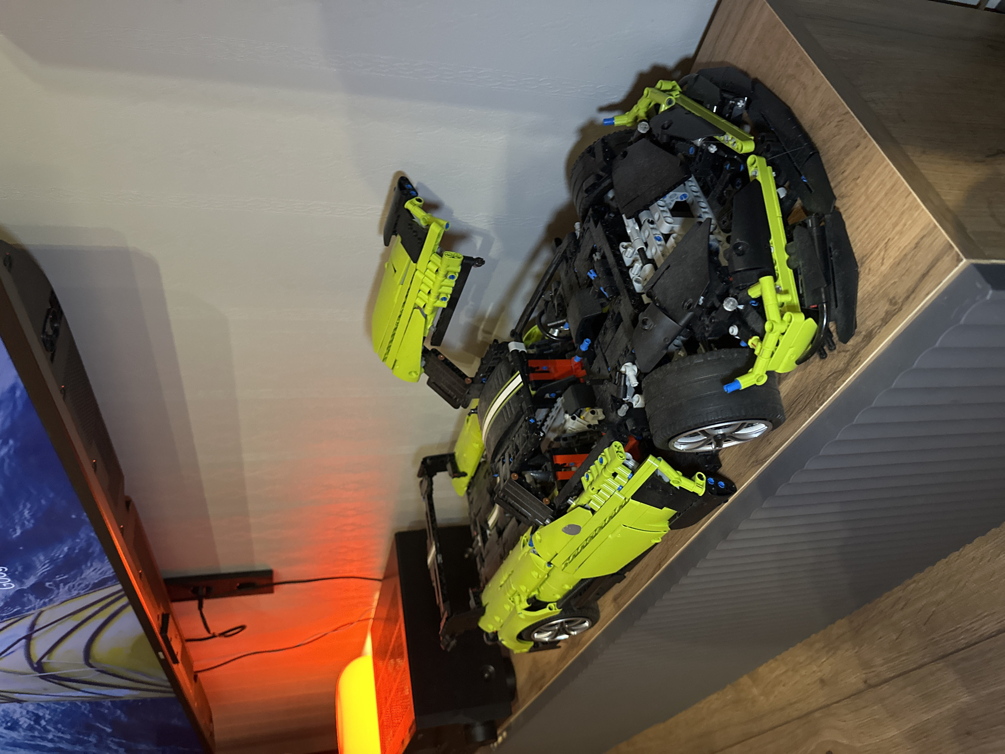 The image shows a partially assembled green and black Lego Technic sports car model sitting on a wooden surface. The model has its front hood open, revealing some of the internal components. To the left of the car is a TV screen displaying a blue image. Below the TV is a small black device with wires and a vertical orange light. The scene appears to be indoors, situated near the wall. The location of the image in the city of Perm, Russia, is not directly inferable.