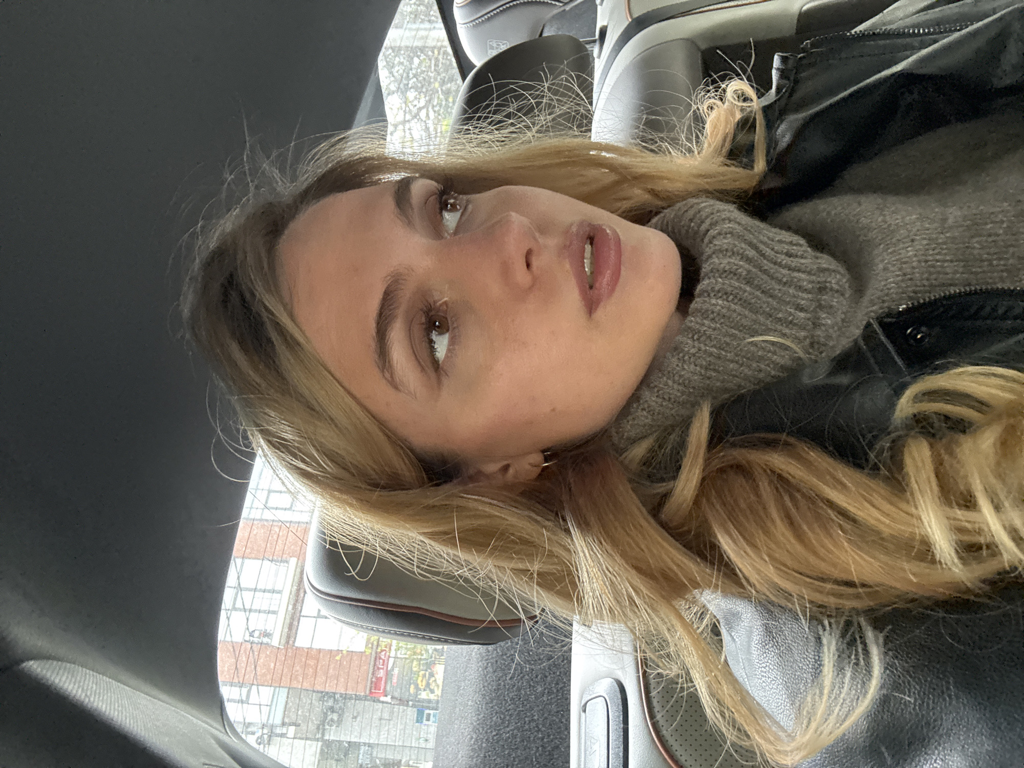 The image is a vertically oriented shot of a woman reclining or lying down inside what appears to be a car. Her body is positioned horizontally within the frame, with her head towards the top left. She has long, wavy blonde hair, fair skin, and light brown eyes, looking upwards and slightly away from the camera with a neutral or contemplative expression. She is wearing a gray turtleneck sweater under a black leather jacket.
The setting is the interior of a modern vehicle, characterized by black leather car seats with tan or brown accents, visible around her and in the background. Bright, natural daylight illuminates the scene, indicating it is daytime. Through the car window, an urban or suburban environment is visible. This includes a brick building with multiple windows and what appear to be bare tree branches, suggesting an autumn or winter season. A red sign, partially displaying the text "SPA", can be discerned through the window in the background. The lighting emphasizes the textures of her clothing and her facial features.