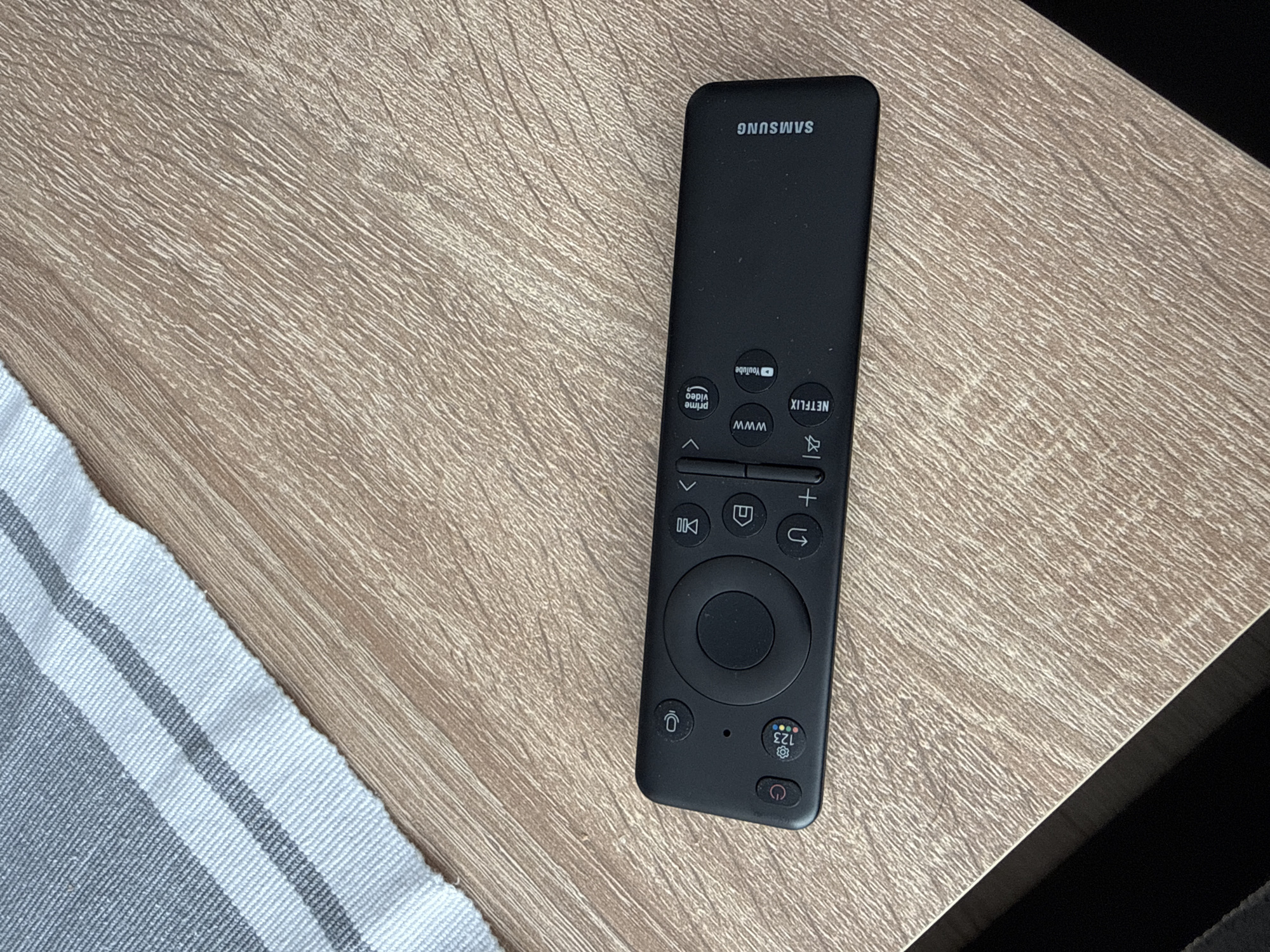The scene is a close-up shot of a Samsung TV remote resting on a light brown wooden surface. A small portion of a striped gray and white fabric is visible on the left side of the frame. The remote is black, and the Samsung logo is visible at the top. The buttons are arranged in a typical remote control layout, including volume control, channel changing, navigation, and dedicated buttons for Netflix, YouTube, and Prime Video. The light source appears to be overhead. There is no indication of this image being in Lovech, Bulgaria.