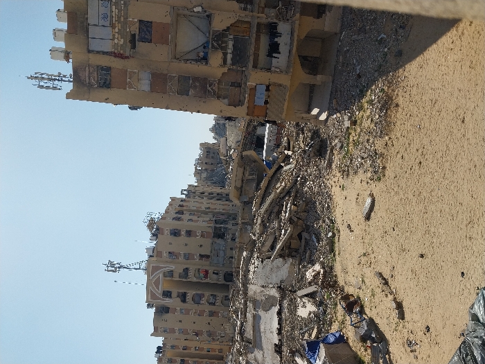 The image depicts a wide view of a severely damaged urban area in Khan Yunis, Palestinian Territory, under a clear blue sky. The foreground is composed of sandy ground with scattered small debris and larger fragments of building materials. A dark, possibly tarp-covered object is situated in the lower-left corner, and a twisted metallic structure rests on the sand in the mid-left. Blue tarps are draped over a pile of rubble near the left edge.
The midground is dominated by a vast field of pulverized concrete, twisted rebar, and general building debris, indicating the extensive demolition of structures. A white vehicle, resembling an ambulance or utility truck, is partially visible amidst this rubble toward the center-right.
Multiple multi-story buildings, constructed of tan-colored concrete, frame the scene. On the left, a cluster of residential-style buildings exhibits widespread damage, including numerous blown-out and boarded-up windows, with communication antennas and cellular towers on their rooftops. The building on the right, positioned closer to the viewer, displays severe facade damage with missing windowpanes, some covered openings, and visible balconies, one with laundry hanging. This building also has multiple communication antennas mounted on its rooftop. No individuals are discernible within the scene.