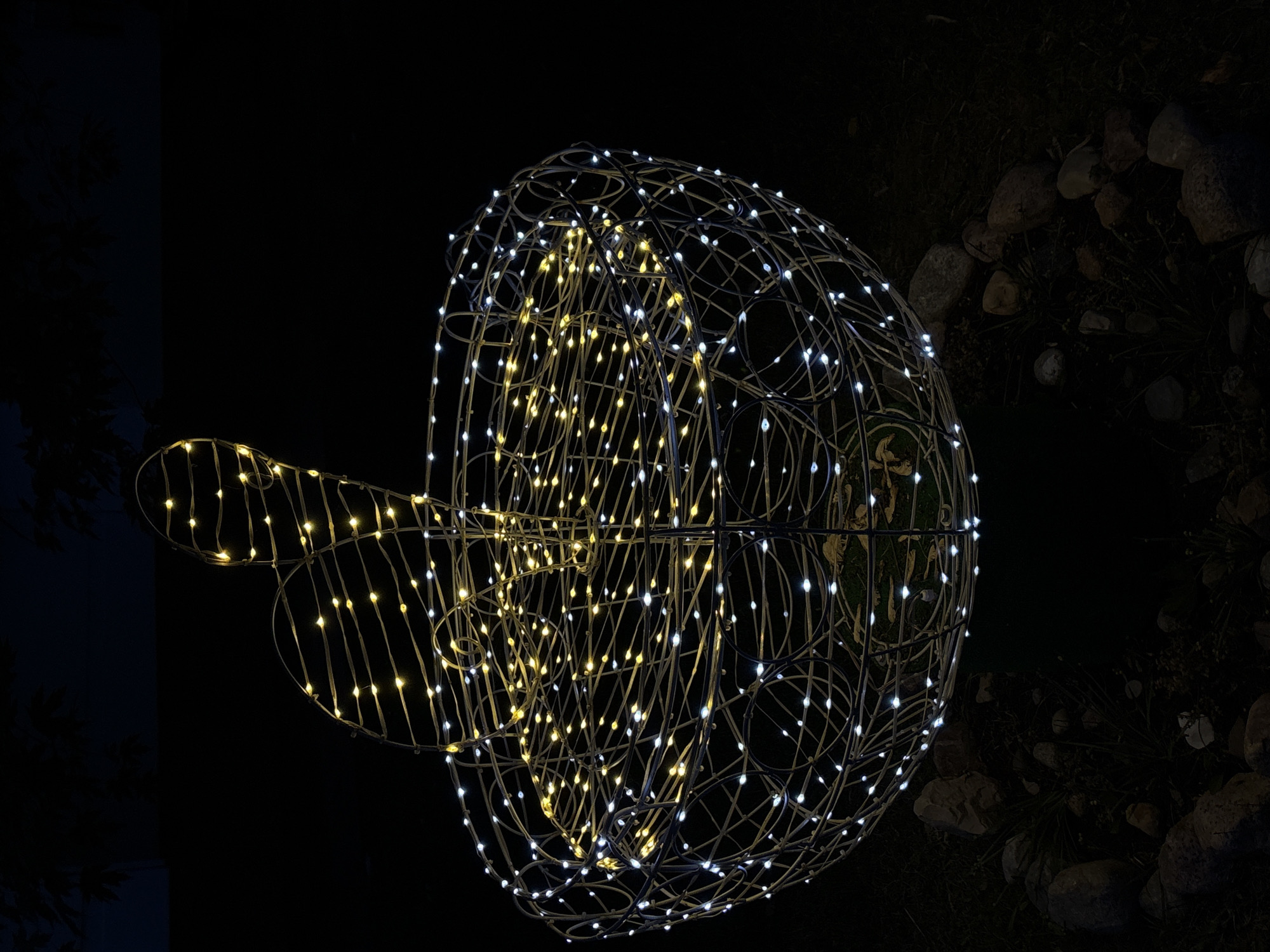 A wireframe sculpture, possibly a Easter bunny or hare, adorned with white and yellow LED lights, is illuminated at night. The sculpture is in a large, roughly spherical basket shape with a separate upright form emerging from it. The basket is illuminated with both white and yellow lights, creating a festive glow. The upright form, also lit with yellow lights, appears to be the head and ears of a rabbit. The sculpture is placed outdoors, with dark foliage visible in the upper background and rocks and grass surrounding its base. The image suggests an outdoor decoration, likely for a holiday or event, situated in a residential or public garden setting. The provided context indicates the image was taken in Vitsebsk, Belarus.