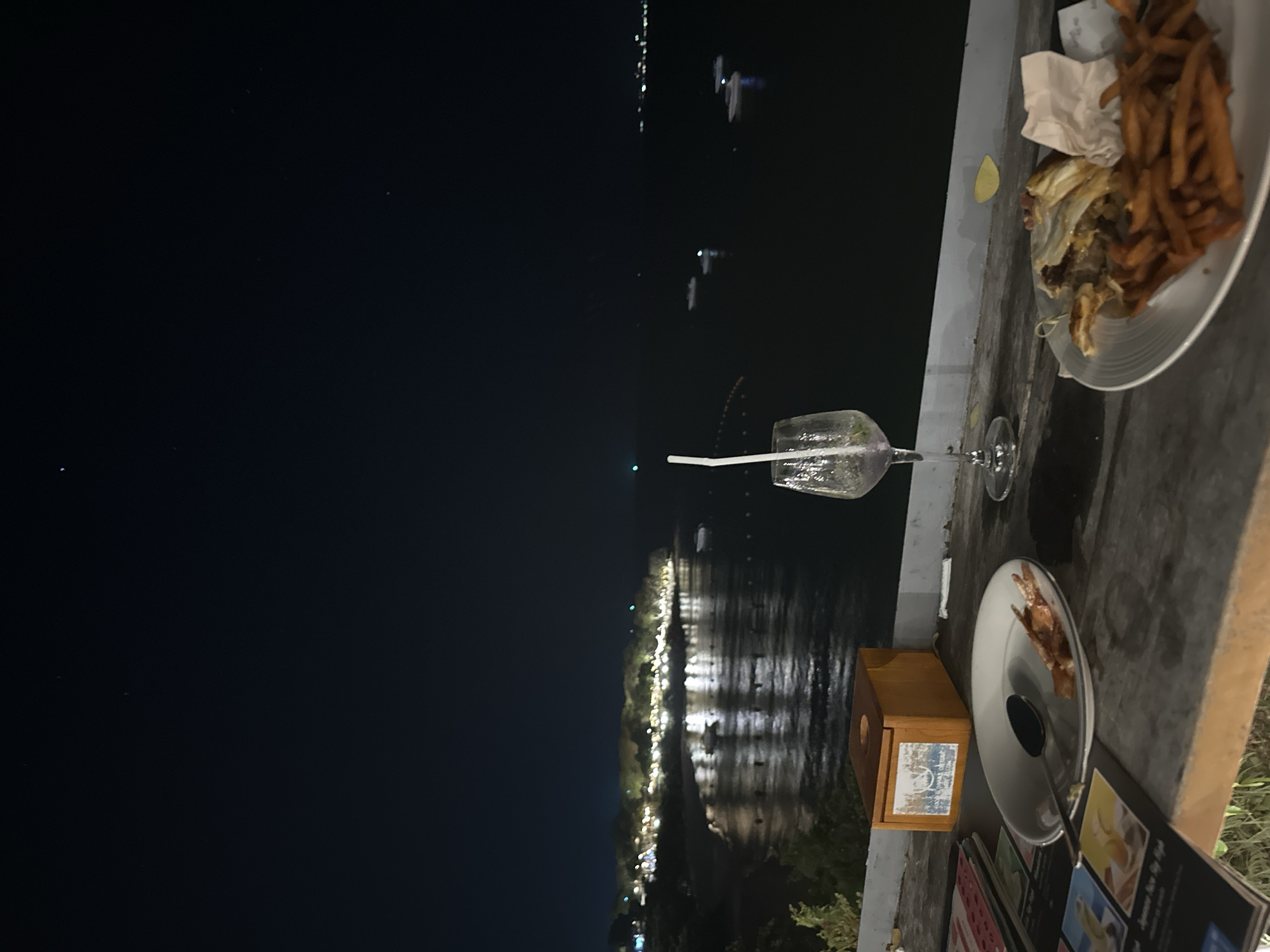 The photograph depicts a nighttime scene at a restaurant in Pattaya City, Thailand. A table overlooks a body of water, presumably the sea, with lights visible along the shore. A half-eaten meal of chicken and fries is on a plate, indicating recent dining activity. A wine glass with a straw also sits on the table. The table surface is visible, with part of a menu and a wooden box-like object that could be a napkin holder. The focus is on the aftermath of a meal and the outdoor setting.