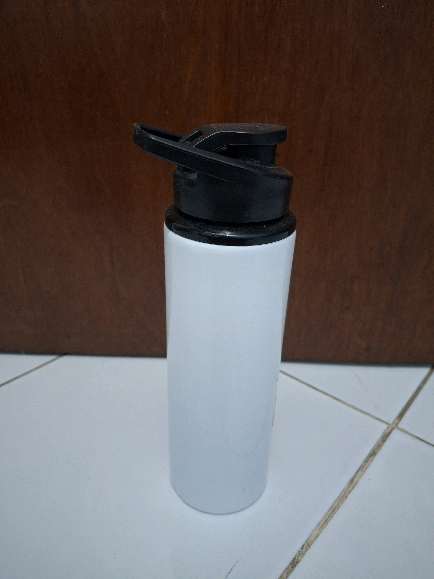 A white water bottle with a black lid is placed on a tiled surface. The background is a dark brown wooden wall. The scene is indoors, possibly in a home or office. There are no people or animals present in the image. The lighting is even and does not cast any harsh shadows. The image appears to be a still life photograph. The location in Lumajang, Indonesia is not discernible from the image itself.