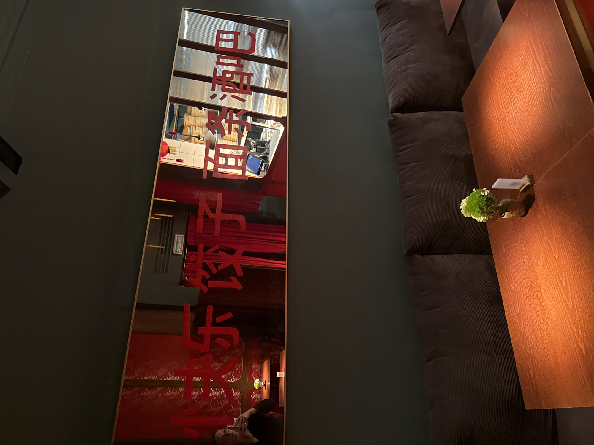 This media file captures an indoor scene, likely within a restaurant, bar, or lounge. The image features a prominent, tall vertical mirror on the left side, adorned with large, red East Asian characters, which appear to be Chinese. These characters are vertically arranged and seem to spell "日照茶坊" (Rìzhào Cháfāng), translating to "Rizhao Tea House" or "Sunshine Tea Workshop."
The mirror reflects the room opposite it, revealing details such as slatted windows or blinds, a counter area with cylindrical items (possibly decorative barrels), a red lantern or light fixture, and a red wall with patterned wallpaper or fabric. A visible vent in the reflection bears the Russian word "ВЫХОД" (VYKHOD), meaning "Exit." In the lower part of the reflection, a person is seen reclining or sitting back, looking at a mobile phone.
To the right of the mirror, a long, dark gray or brown upholstered sofa with a ribbed texture provides seating. In front of the sofa is a reddish-brown wooden table. On the table, a small green decorative plant, possibly moss or a small succulent, sits in a container alongside a small, white rectangular object.
The setting suggests an establishment with an East Asian theme, located in Moscow, Russia, as indicated by the context and the Russian "Exit" sign. The lighting is dim, creating a relaxed and intimate atmosphere, characteristic of an evening or night setting. The activity shows an individual in a state of leisure, engaging with their phone.