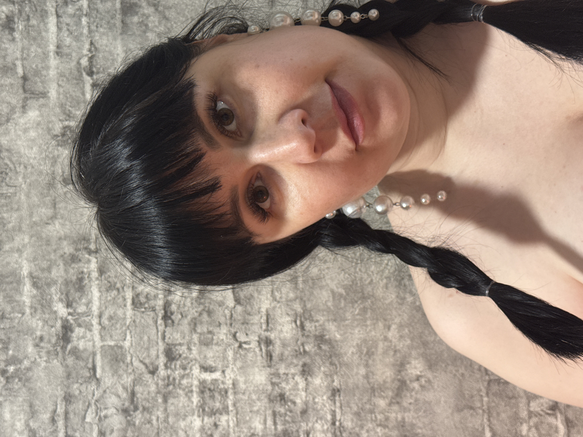 A woman with fair skin, dark brown eyes, and dark hair styled in braids with blunt bangs is depicted in a close-up portrait against a gray textured background resembling a brick wall. She is wearing a pearl necklace. The image is rotated counter-clockwise. The location in the city of Zav'yalovo, Russia is unknown.
