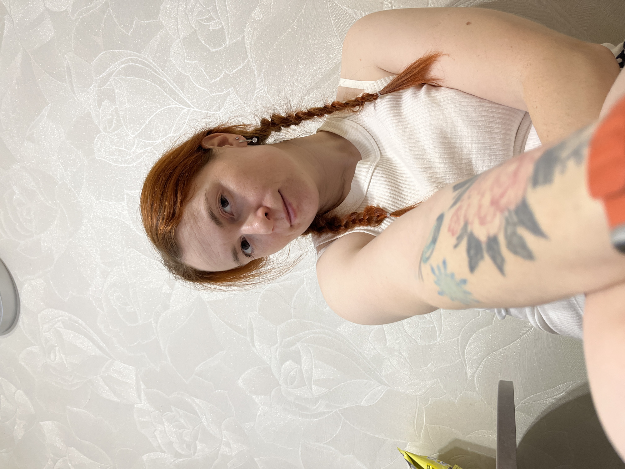 A woman with red hair braided into two plaits is photographed indoors. She is wearing a white tank top and has tattoos on her left arm. The background is a wall with a subtle floral pattern. A small portion of an object with a yellow and green package is visible in the bottom left corner. A reflective metallic object is near the bottom right corner. The photograph was taken inside a space in the city of Chaikovsky, Russia.