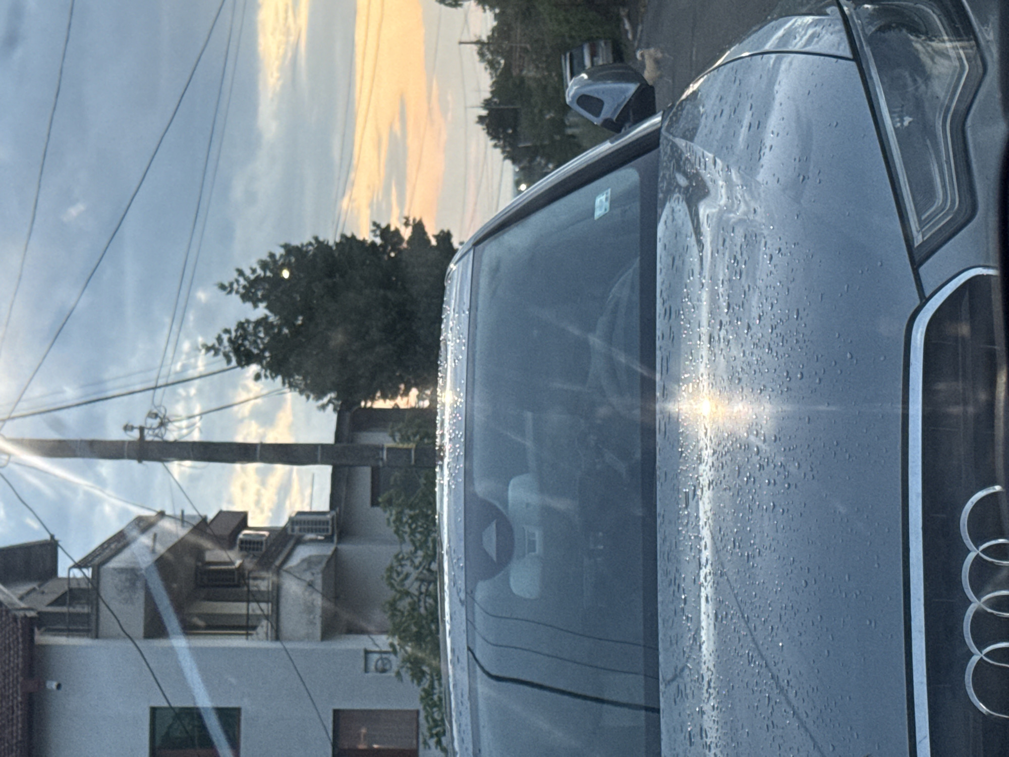The image, rotated 90 degrees clockwise, shows a close-up of the front of a silver Audi car. Water droplets cover the hood. The Audi logo is visible. The car is in an urban environment, likely in Burgas, Bulgaria, as indicated in the prompt. Reflected in the windshield are buildings, trees, power lines, and a sky with clouds and a sunset. One building has an exterior air conditioning unit. A black object is mounted above the front passenger side window. The scene appears to be early morning or late evening, indicated by the soft light.