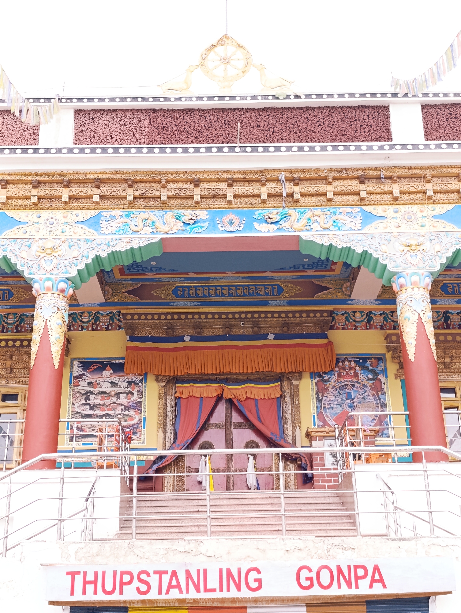 The image shows the entrance to Thuptstanling Gonpa, a Buddhist monastery in Leh, India. The entrance features an elaborate archway with intricate carvings and paintings, characteristic of Tibetan Buddhist architecture. Red columns support the arch, and colorful prayer flags are visible at the top of the structure. Steps lead up to the entrance, and railings provide support for visitors. Two paintings adorn the walls flanking the entrance, one depicting a procession with elephants and another a Wheel of Life, a central symbol in Buddhism. A sign in red lettering at the bottom of the image reads "THUPSTANLING GONPA". The overall scene suggests a religious and cultural landmark with vibrant artistic details.