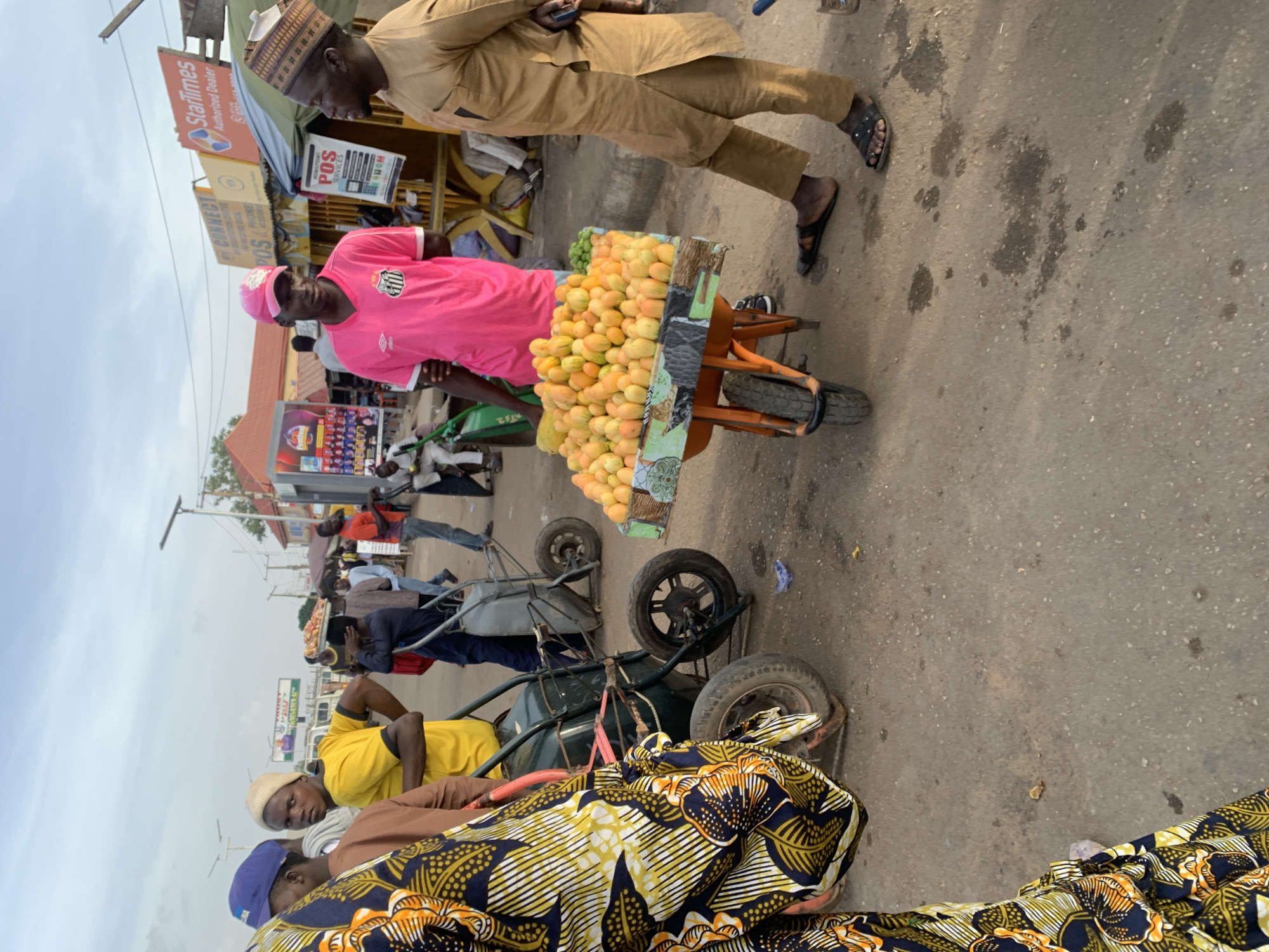 The image depicts a street scene in Gura Top, Nigeria. A man wearing a pink shirt and matching pink baseball cap is standing beside a wheelbarrow overflowing with mangoes, suggesting he is a vendor. Another man in a brown suit and cap is standing beside the mango vendor. There are several other individuals present on the street, and many of them are standing alongside other wheelbarrows. The background includes storefronts with signs, one of which reads "Startimes Authorized Dealer". The weather appears overcast. The road is asphalt, with visible stains and discoloration.