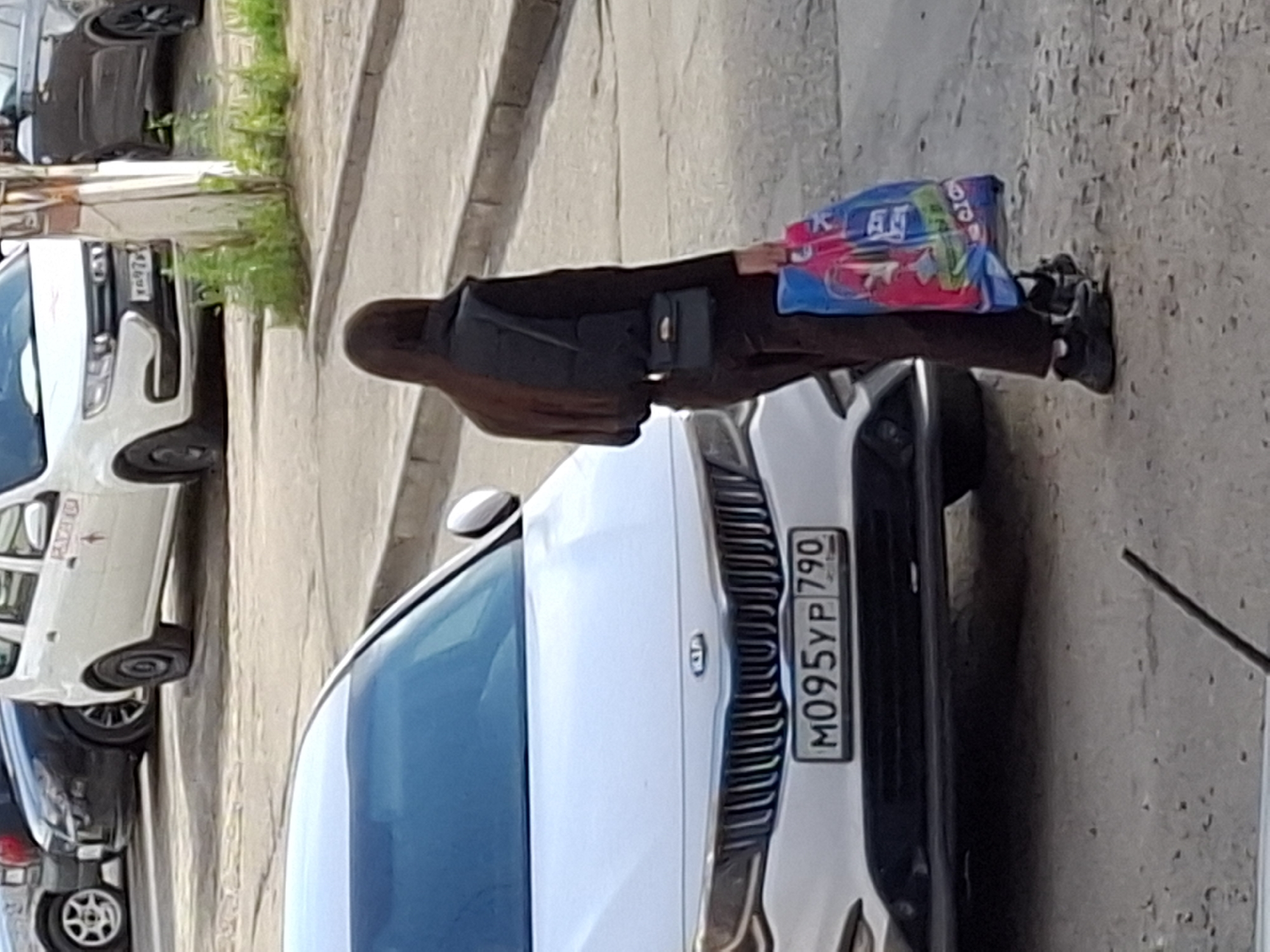 A woman with long brown hair, wearing a dark vest over a dark top and dark pants, and black sneakers, walks away from the camera carrying two colorful shopping bags in her right hand. She also has a small black handbag attached to her belt. She is walking on a paved surface next to a white Kia sedan. The car has a license plate that reads "M095YP 790". In the background, other vehicles are parked on the street, including a white van and a dark-colored car. The environment appears to be an urban street with a sidewalk and vegetation growing along the curb.