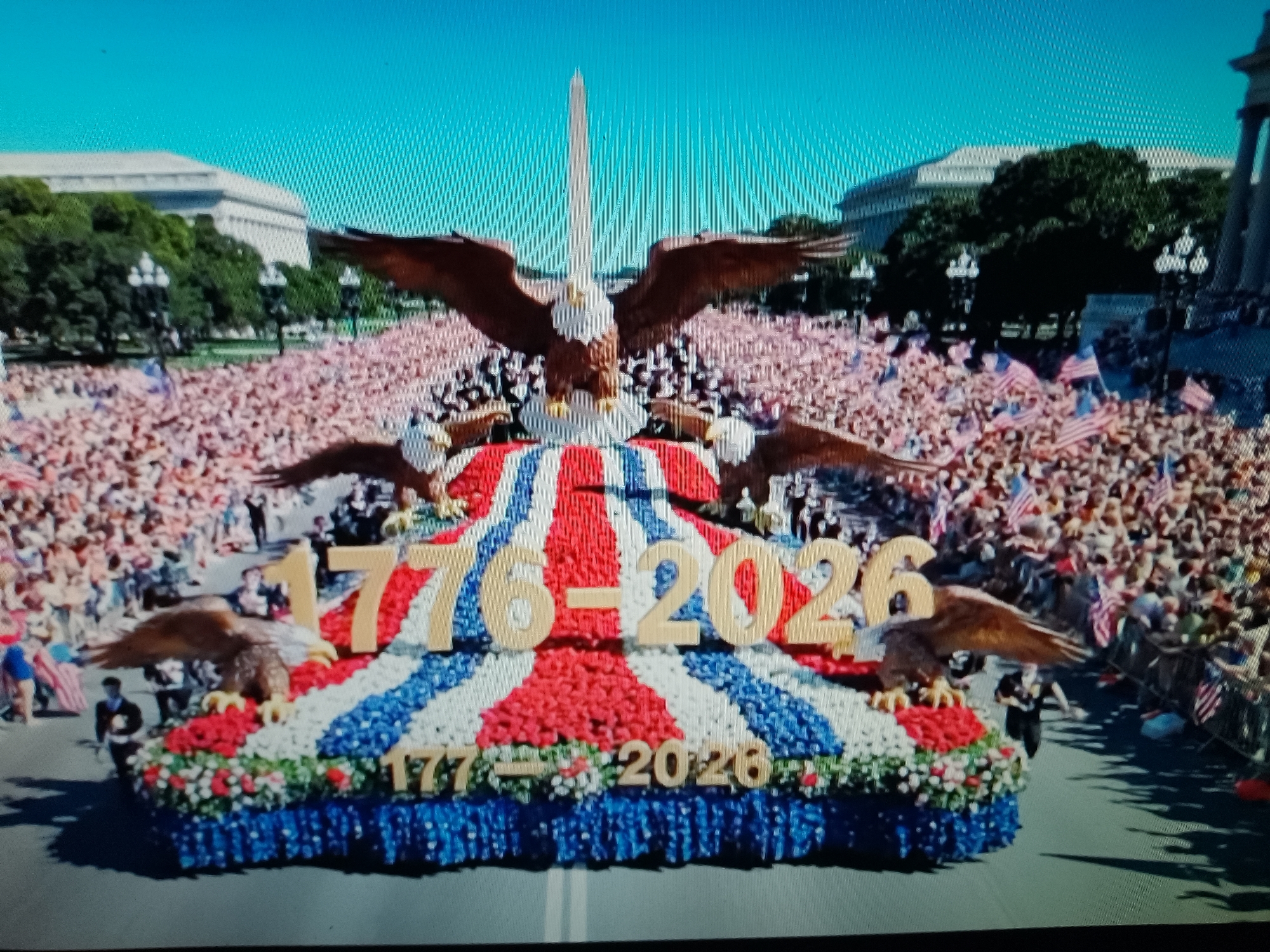 The image depicts a patriotic parade float decorated with red, white, and blue flowers and featuring a large eagle statue with outstretched wings. The number "1776-2026" is prominently displayed on the float, along with a smaller inscription of "1776 - 2026" at the base. A crowd of people, many holding American flags, are visible in the background, suggesting a national celebration. The Washington Monument can be seen in the distance. The scene is set in a public space, possibly a city street, with buildings and trees lining the route. However, the location is not in Chiajna, Romania.