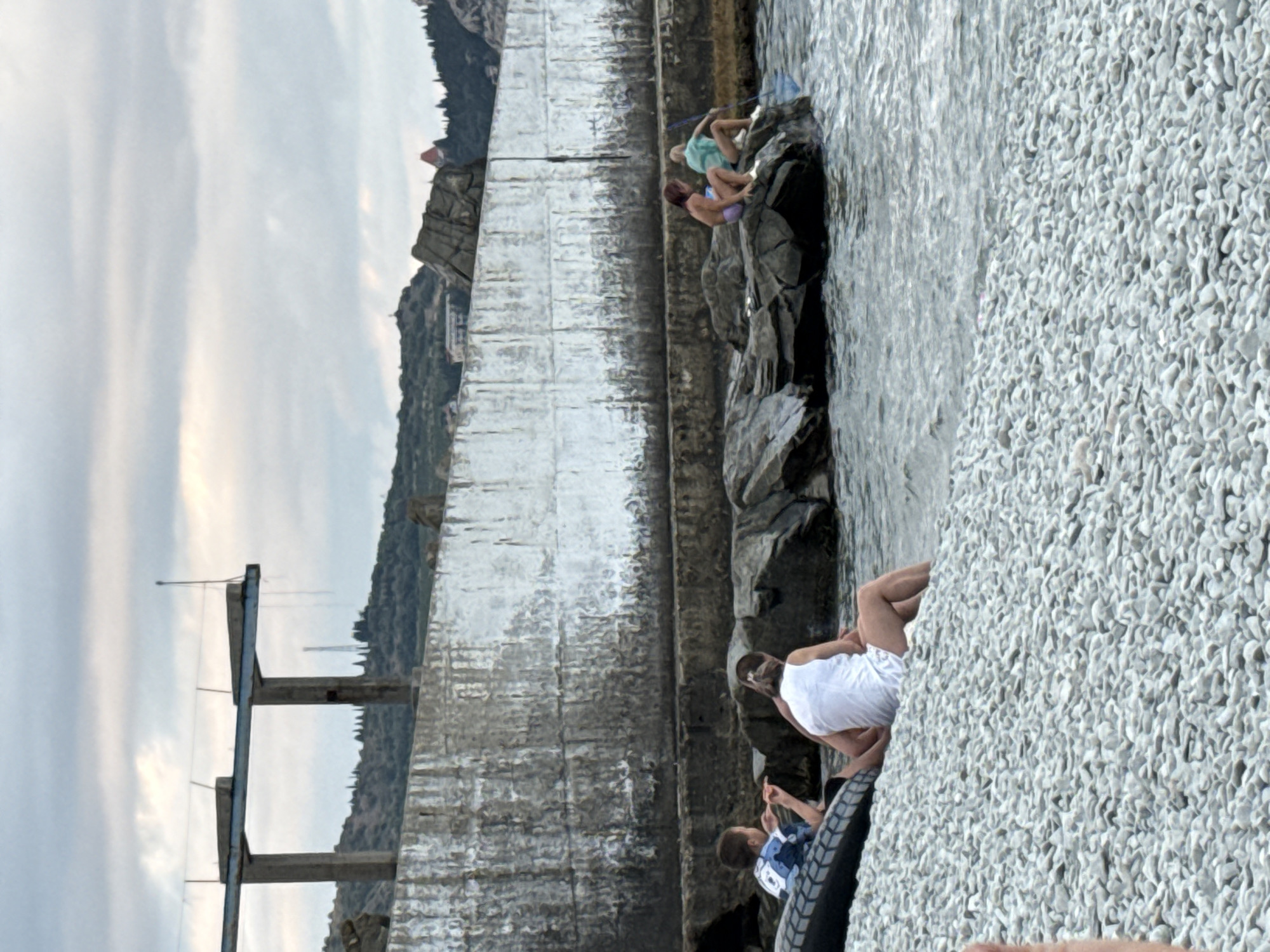 The image depicts a rocky shoreline in Partenit, Ukraine, adjacent to a large, weathered concrete wall. In the foreground, a child in a blue and white shirt sits on the pebble beach, next to a black inflatable tire. Nearby, a person in a white top and shorts is seated on the pebbles, facing away from the camera.
Further out, several people are positioned on rocks at the water's edge. Two individuals, one in a purple bikini and another in a blue top, are seated on the rocks. Another individual, wearing a light blue top and shorts, is also on the rocks, holding a fishing net. The sea is calm, with gentle waves lapping against the rocks and shore.
Behind the concrete wall, a hilly landscape is visible, covered in green vegetation and scattered trees. The sky is overcast with clouds. Overhead, a concrete structure with two pillars supports a horizontal beam, likely part of a disused pier or diving platform, with thin wires extending from it. The overall scene suggests a relaxed atmosphere by the sea, with people engaging in leisure activities.