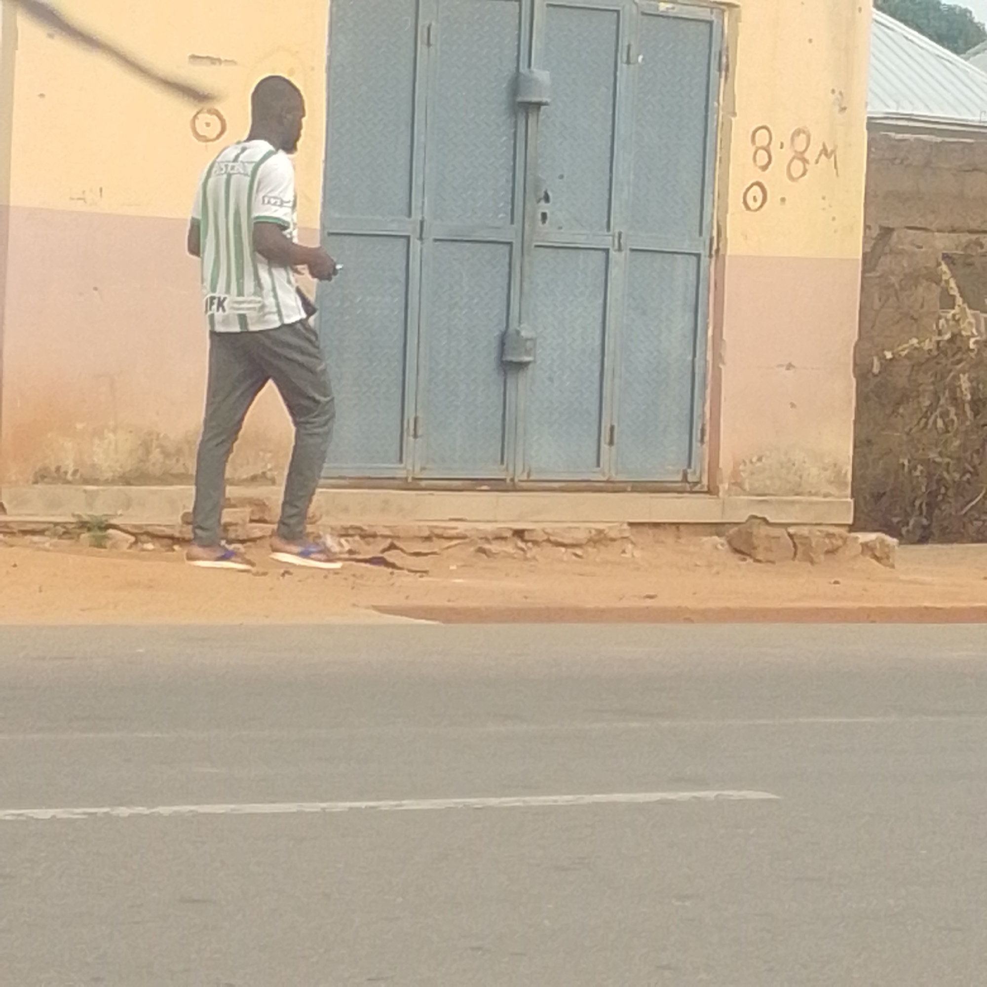 This media file captures a full-body shot of a man walking along a sandy path next to a building with a large metal door. The man is wearing a white t-shirt with green stripes and dark gray pants, and he appears to be holding a small object in his right hand. He is wearing sandals.
The building has a light yellow and pink facade, with some faded markings. On the wall to the right of the door, there is handwritten text that appears to read "8.8M" and a circular symbol with a dot in the middle. The metal door is large and appears to be made of corrugated metal, divided into four sections.
The scene suggests an outdoor setting in what seems to be a semi-arid environment, given the sandy path and the dry vegetation visible behind the building on the right. The lighting indicates it is daytime, but the specific time is not clear. There are no obvious cues about the weather, though the dry ground might suggest it's not currently raining. The overall mood is neutral, with the man simply walking.
The location context provided is Gombe, Nigeria, which could align with the visual cues of the environment. The man's attire and the style of the building are consistent with a location in West Africa. The image is captured from a low angle, looking up slightly at the man and the building, with a road visible in the foreground.