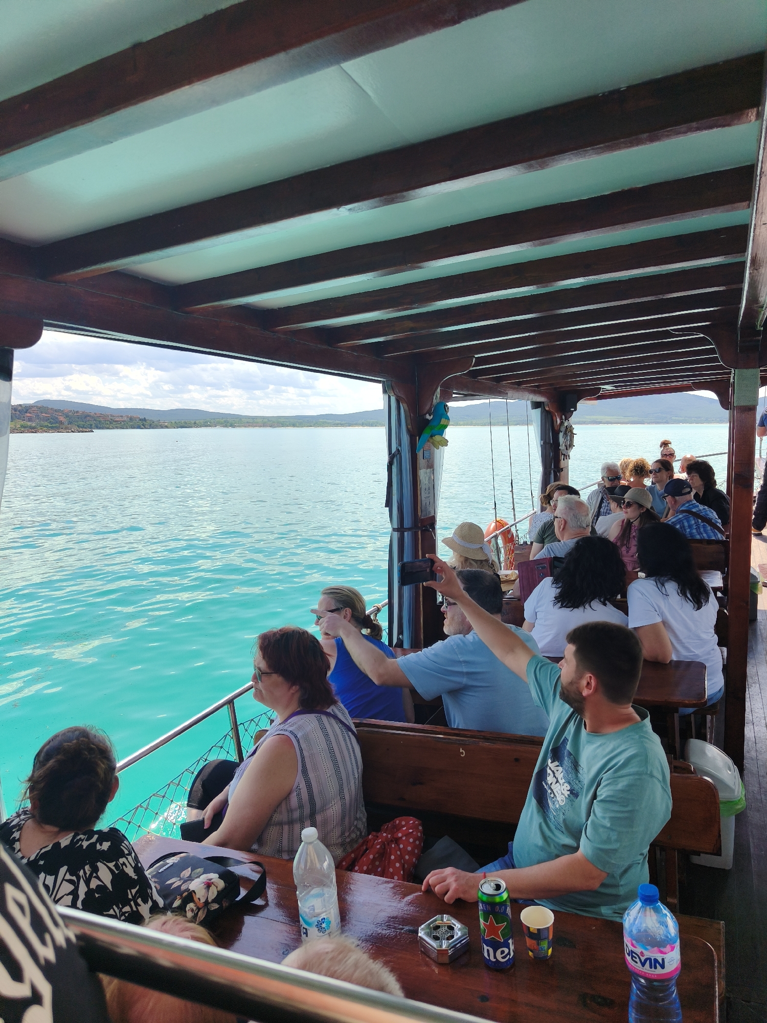 The image shows a group of tourists on a boat, likely during a tour in Sozopol, Bulgaria. The water is turquoise, and land is visible in the distance. The boat is covered with a wooden roof structure. People are seated on wooden benches and appear to be enjoying the view. One man is taking a photo with his phone. On a table, there is a bottle of water, a can of Heineken 0.0, a paper cup, and an ashtray.
