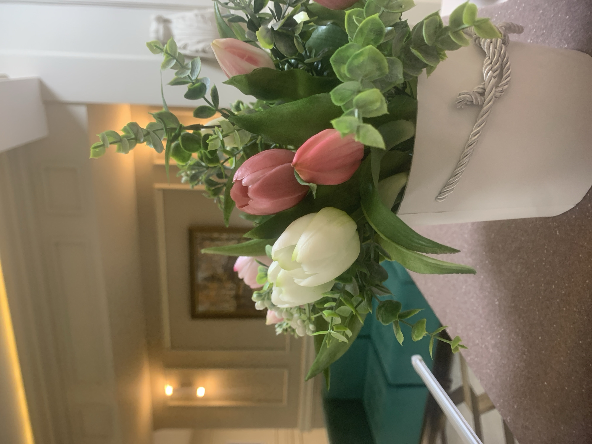 The image shows a floral arrangement in a white container with a twisted rope handle, featuring tulips in pink, white, and red hues, interspersed with green foliage. The arrangement sits on a brown surface, potentially a table. The background reveals a blurred interior, suggesting a room with light-colored walls, decorative molding, and a framed picture. In the background are some lights, and a partially visible teal-colored piece of furniture, such as a sofa. The location is not discernible from the image to be Izhevsk, Russia.
