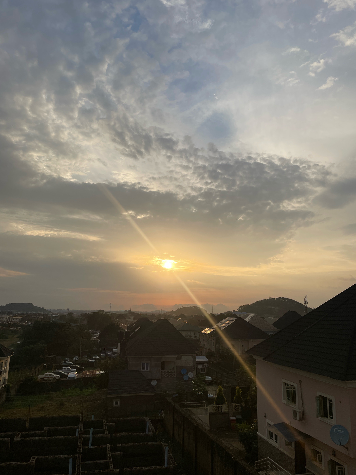 This is an aerial view of a residential neighborhood in Abuja, Nigeria, during a sunset. The sky is filled with dramatic clouds, with the setting sun casting a warm golden glow across the scene. A prominent sunbeam cuts diagonally from the upper left to the lower right. In the foreground, the rooftops of houses are visible, with one light pink house on the right featuring windows and an air conditioning unit. Further into the scene, more houses with dark roofs are clustered together. A dirt road or unpaved area is visible, with parked cars, including white sedans and vans. In the middle ground, construction is underway, with the brick foundations of several buildings laid out. Beyond the immediate residential area, rolling hills and more distant buildings can be seen against the hazy horizon. The overall impression is one of a developing urban landscape bathed in the soft light of sunset.