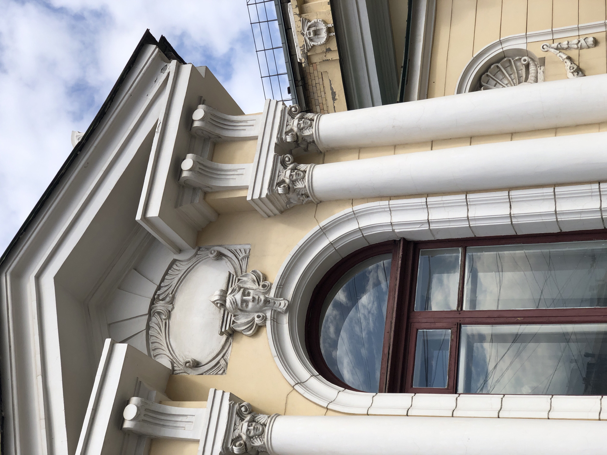 This image captures a close-up, upward-tilted view of the ornate facade of a building in Krasnoyarsk, Russia. The building features a pale yellow wall adorned with extensive white architectural details, suggesting a classical or neoclassical revival style.
The main subjects are the building's decorative elements. Prominently featured are two large, cylindrical white columns or pilasters with elaborate capitals carved with human faces, located on the upper part of the facade. Below a round, dark-framed window, another column capital with a sculpted face is visible. Next to the round window, a decorative element shaped like a stylized face with flowing hair or foliage is affixed to the wall. Further decorative motifs include ornate oval frames, shell designs, and volutes. The roofline shows white cornices and a dark, weathered edge with some peeling paint.
The scene is set outdoors during the daytime. The weather appears partly cloudy, with blue sky and white clouds visible in the background and reflected in the building's windows. The round window, with its dark brown frame, reflects the sky, while a rectangular window below it reflects both the sky and what appear to be overhead wires or tree branches. The building's white plasterwork shows minor cracks and weathering, indicative of age. No specific activity is taking place, as it is a static architectural shot. There is no visible text in the image.