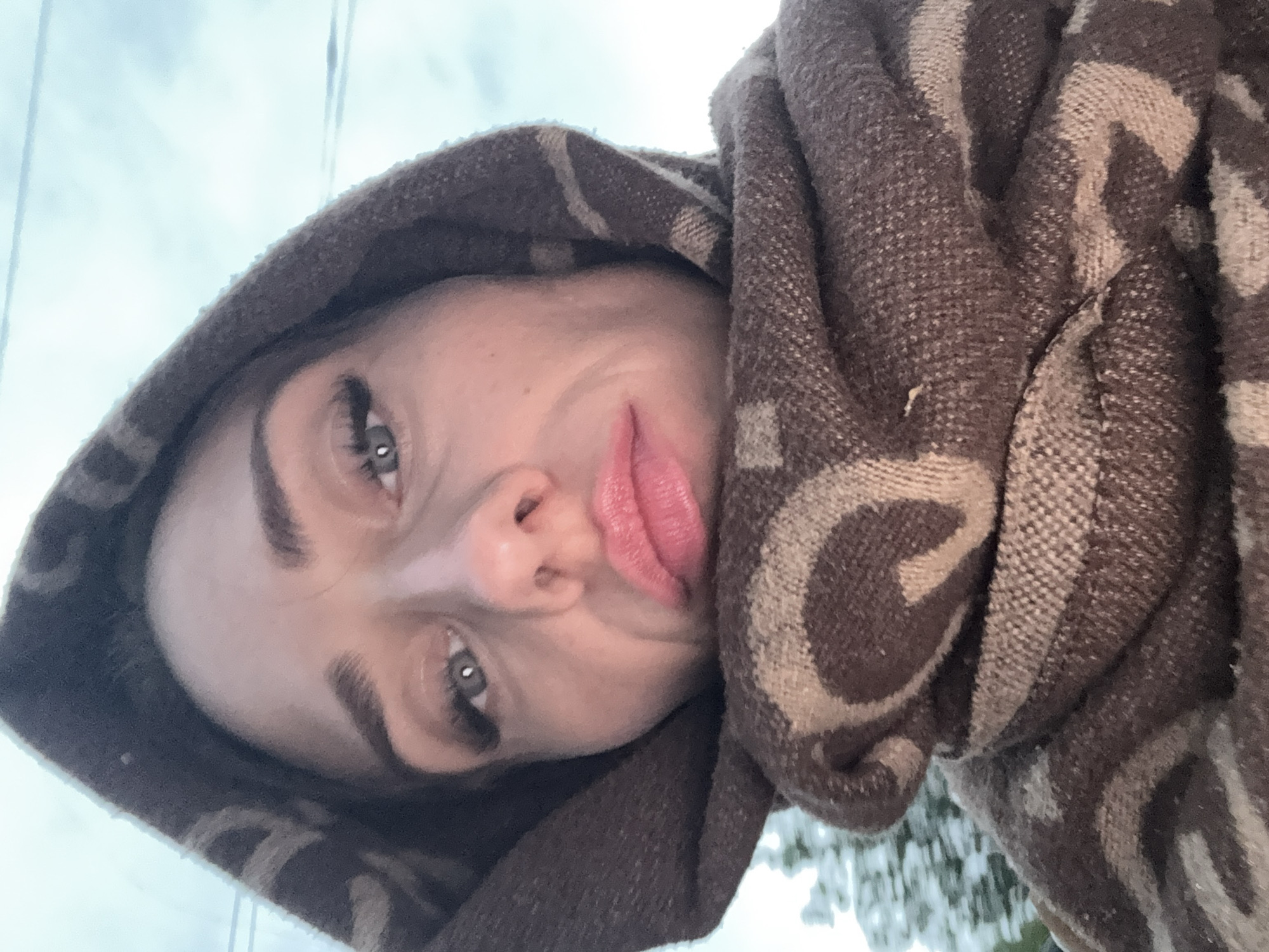 The image is a close-up selfie of a woman wearing a brown hooded garment with a patterned scarf wrapped around her neck and face. She has visible eyelashes and pink lipstick. The background shows a blurred, overcast sky and some out-of-focus greenery, suggesting an outdoor setting. No specific landmarks or identifiable features of Saint Petersburg are visible in the image.