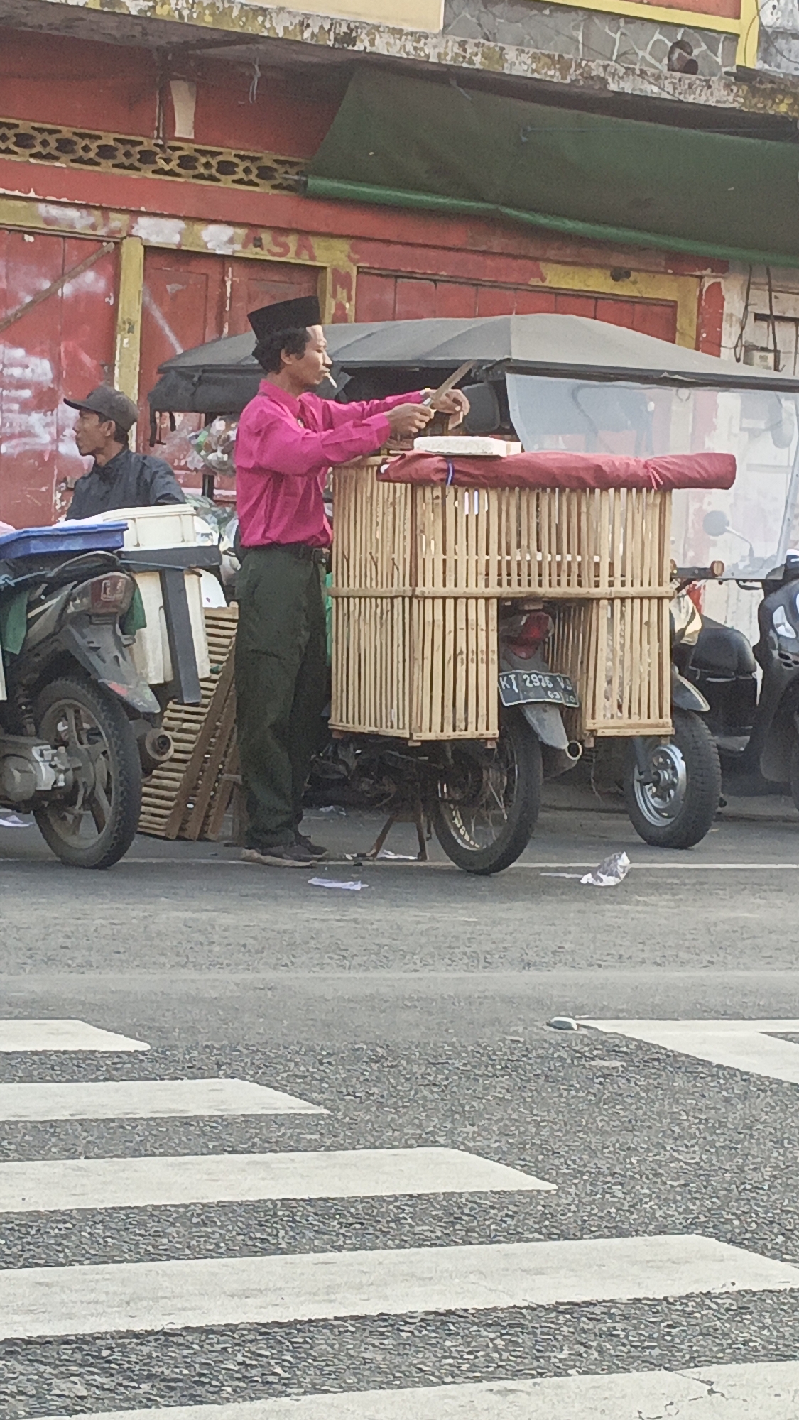 A man wearing a pink shirt and a black cap stands next to a motorcycle with a bamboo cage attached to it. He is holding a knife and appears to be preparing food or goods on a white surface. Another man in a dark shirt and a cap sits on another motorcycle to the left. The scene is on a street in Srono, Indonesia, with buildings and other motorcycles visible in the background. There is a pedestrian crossing in the foreground.