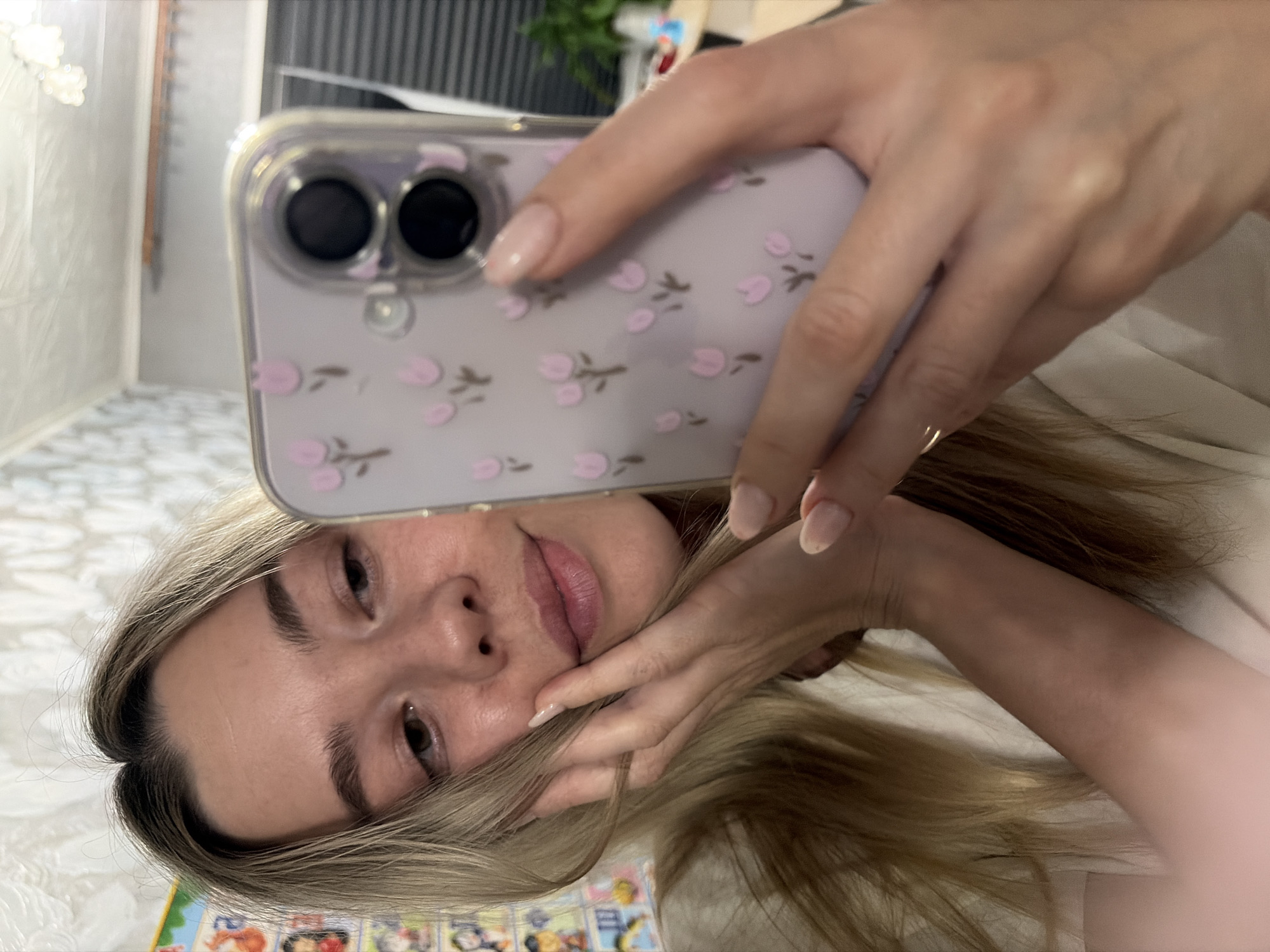 A woman is taking a selfie with her smartphone. The phone has a clear case with a floral pattern. She holds the phone in her right hand, her fingers visible on the screen and along the side of the phone. Her face is partially obscured by the phone, revealing her eyes, nose, and mouth. She has blonde hair that falls across her face and arm. The background includes a patterned floor and a blurred view of a room with blinds. The exact location in Chaikovsky, Russia, cannot be determined from the image.
