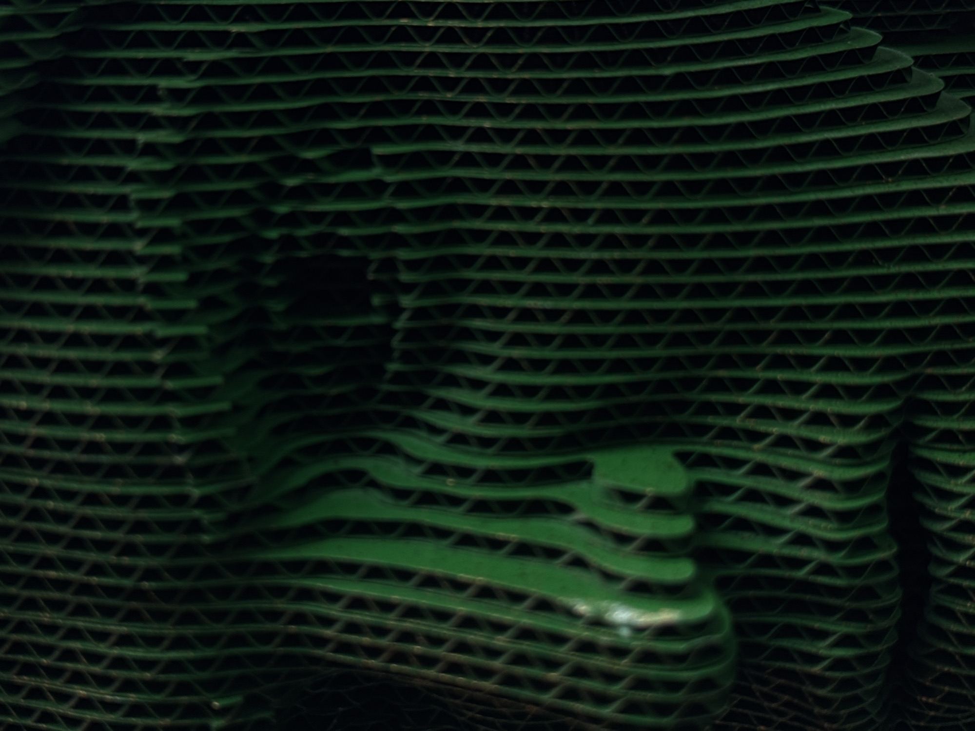 This is a close-up, abstract image of stacked, green corrugated cardboard. The layers of cardboard are cut and arranged to create a topography of curves and hollows, resembling rolling hills or undulating landforms. The texture of the cardboard, with its characteristic triangular ridges and valleys, is prominent throughout the frame. Light and shadow play across the surface, highlighting the dimensional relief of the stacked material and creating a sense of depth. The color is a deep, muted green, which contributes to the organic, landscape-like feel of the composition. The image is abstract, focusing on form, texture, and light rather than any recognizable objects or figures. There is no visible text, indication of time of day, weather, or specific location cues, though the prompt specifies a context of Moscow, Russia. The activity or event taking place is not discernible; the media appears to be a still life or artistic arrangement of materials.
