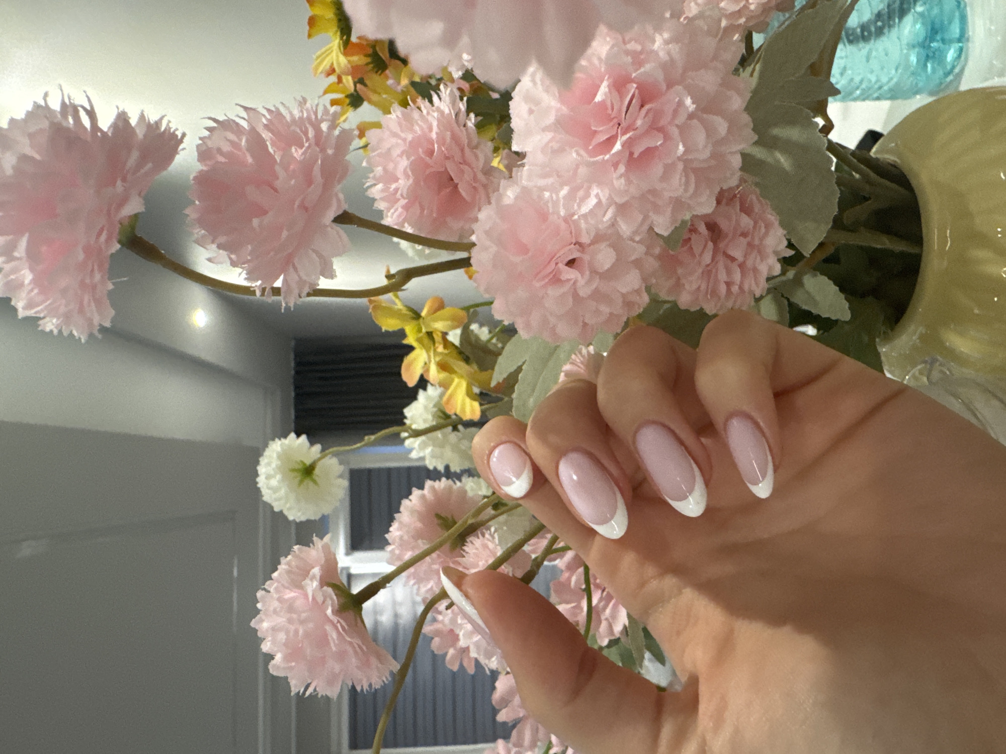 A close-up shot presents a hand with manicured nails featuring a classic French manicure design, with a soft pink base and a crisp white tip. The hand is positioned amidst a bouquet of pink and yellow flowers, which are the main background elements. The flowers appear to be artificial, with detailed petals and varying shades. The lighting in the scene is diffused, creating soft shadows and highlights on the hand and flowers. A glimpse of a room interior, including a window and a light fixture, is visible in the background. There is no specific indication that this image was taken in Burgas, Bulgaria. The scene's composition focuses on the aesthetic of the manicure and the floral arrangement.