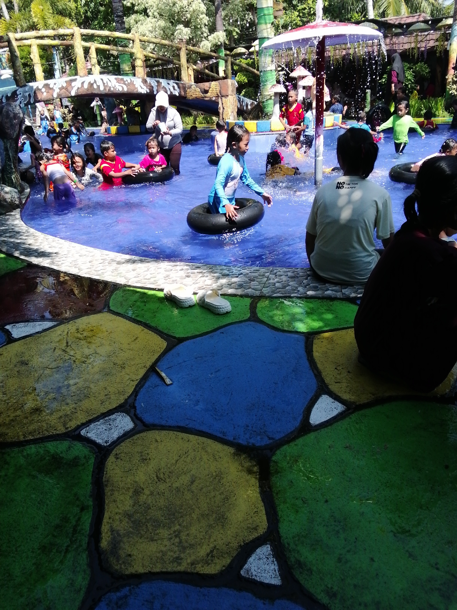 The image depicts a lively scene at a water park, likely located in Tegalwangi, Indonesia, given the lush tropical foliage visible in the background and the style of the park. The central focus is a swimming pool filled with clear blue water, teeming with children and a few adults. Several children are actively engaged in water play, some using inflatable inner tubes. One child, wearing a blue swimsuit, is partially submerged in an inner tube. Another child in a pink top is being held by an adult. A child in a red shirt is also visible in the water. The surrounding area of the pool features a colorful, mosaic-like concrete floor with large, irregularly shaped sections painted in vibrant yellow, green, and blue. The upper portion of the image reveals a wooden bridge structure, likely part of the park's attractions, with tropical trees and foliage behind it. White sandals are placed on the edge of the pool, near the colorful flooring. The overall atmosphere suggests a recreational and family-friendly environment.