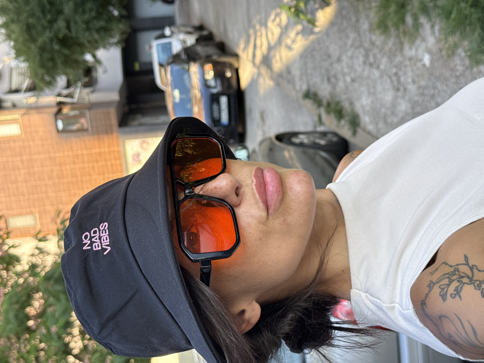 A woman stands on a street in Pleven, Bulgaria, wearing a black bucket hat with the words "NO BAD VIBES" embroidered in pink. She is also wearing oversized square sunglasses with orange lenses and a white sleeveless top. She has dark hair pulled back and a tattoo on her left shoulder. The background shows a street lined with parked cars and buildings. The sunlight casts shadows on the ground.