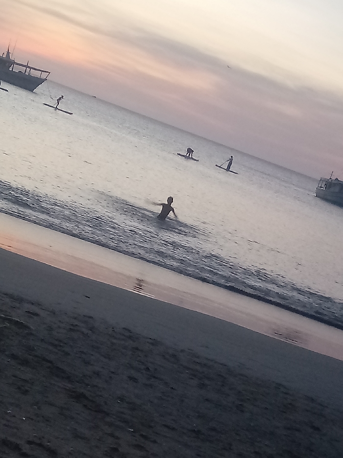 A wide shot captures a beach scene at dusk. In the foreground, dark sand meets the water's edge where a person stands. Further out, several individuals are paddleboarding in the calm sea. Two boats are anchored in the distance on opposite sides of the image. The sky is a soft gradient of pink and light orange, indicating either sunrise or sunset.