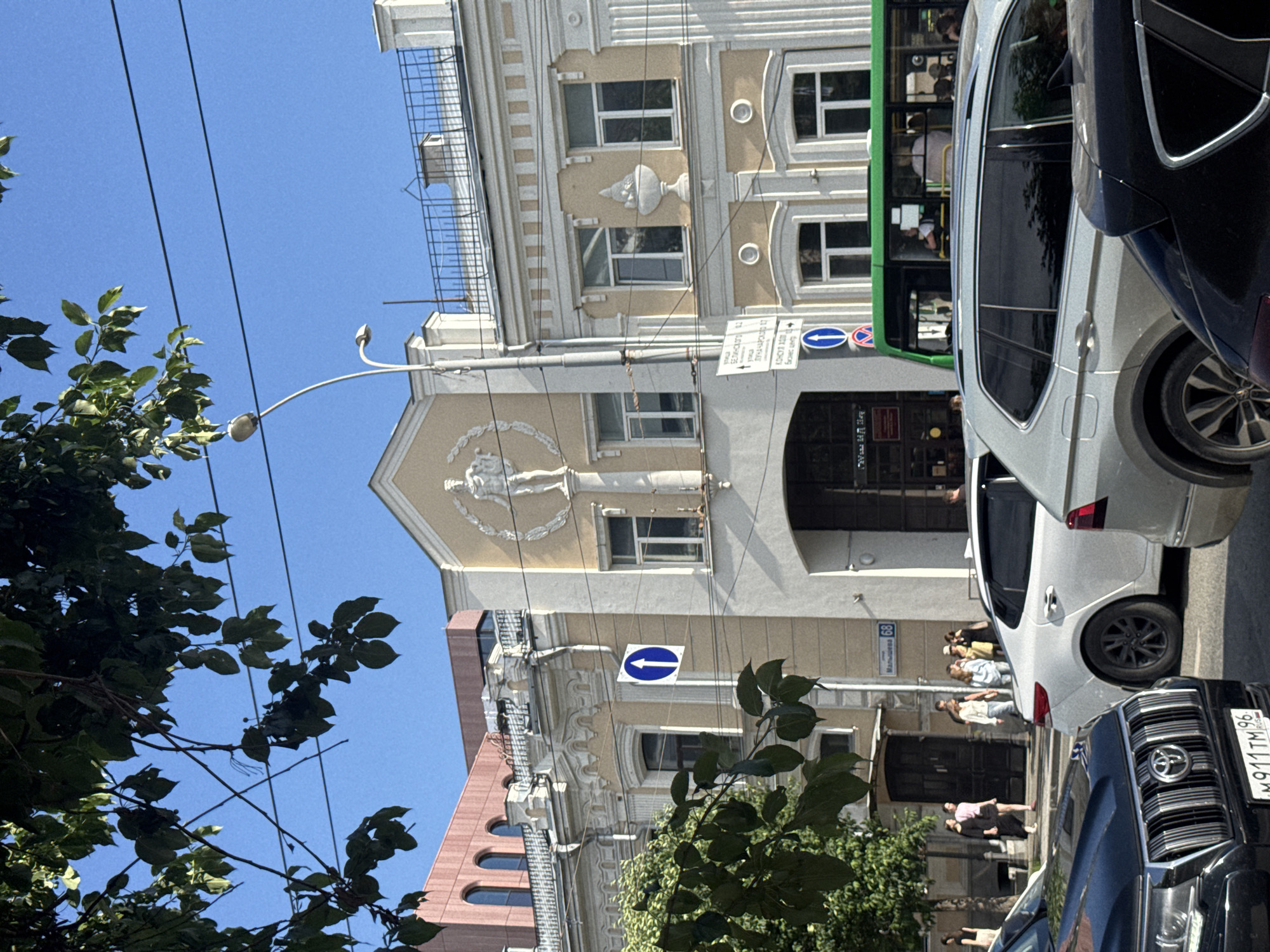 This is a vertical, eye-level, outdoor shot of a street scene in Yekaterinburg, Russia. The image is oriented with the sky at the top and the ground at the bottom. The focus is on the architecture of buildings and parked vehicles, with some foliage in the foreground.
The buildings appear to be older, with decorative facades, arched windows, and balconies. One building has a statue or relief figure above an entrance. A street sign with Cyrillic text and the number "68" is visible.
Several cars are parked along the street, including a white SUV with its rear door open, and a dark SUV in the foreground. A green bus is also visible, with people inside. A few individuals are seen walking on the sidewalk and near the entrance of a building.
The scene appears to be taking place during the day, under a clear blue sky, suggesting fair weather. The overall impression is of a relatively quiet urban street. The text visible includes the street sign and some lettering on the license plate of the dark SUV which reads "M911TM 96".