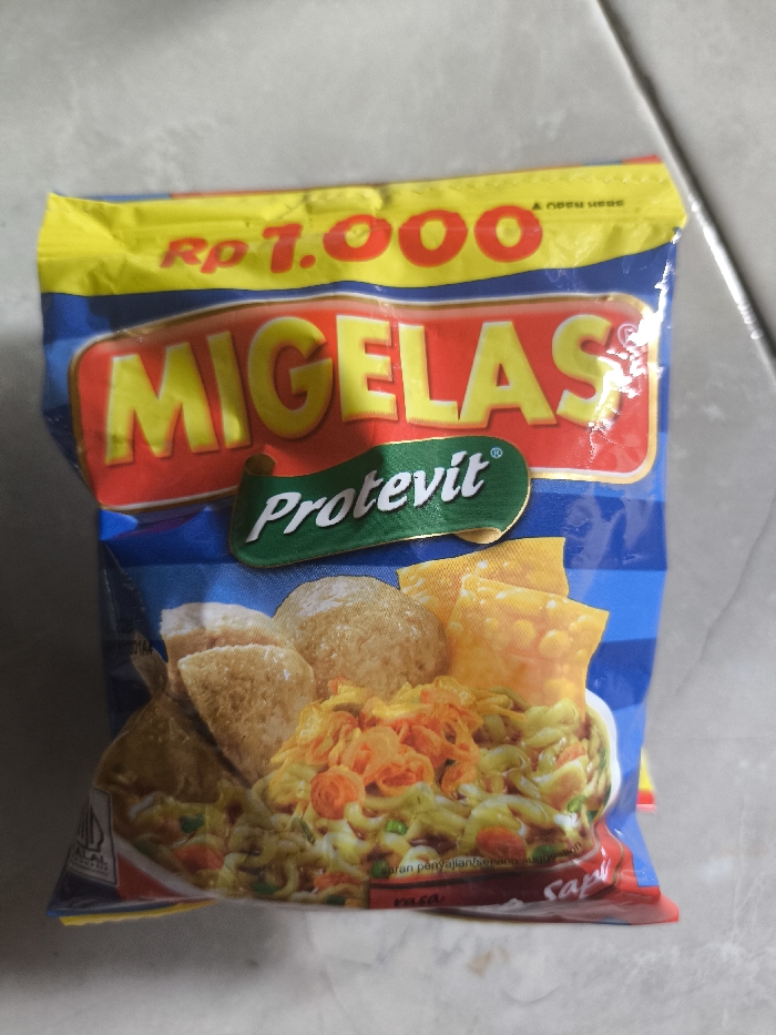 A blue and yellow Migelas noodle package is featured. The words "Rp 1.000" appear at the top of the packaging. A red box with yellow "MIGELAS" lettering sits below. A green banner with "Protevit" in white is next. The bottom half of the package showcases an image of the prepared noodle dish, complete with fried wonton wrappers and meatballs. "Sapi" is written near the bottom edge.