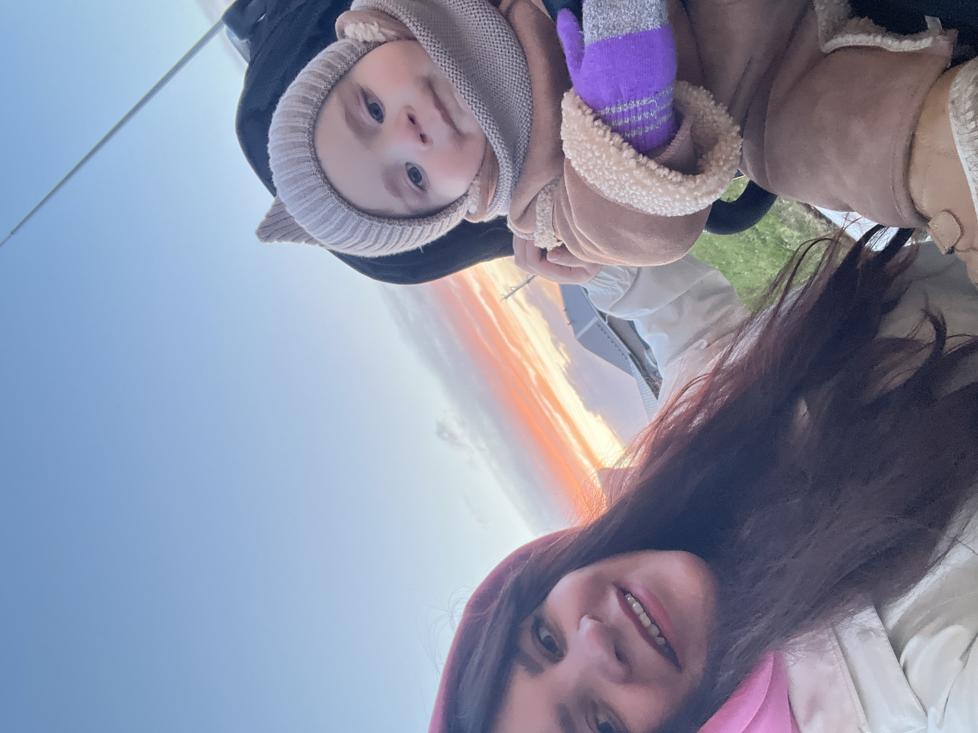 An adult female and a baby/toddler pose for a low-angle selfie, looking up into the camera outdoors in Noviy, Russia. The adult female, positioned on the right side of the frame, has long, dark brown hair and is smiling warmly. She wears a dark pink top or scarf and a light-colored jacket.
The baby/toddler, held by the adult and occupying the left side of the frame, is dressed for cool weather. They wear a light beige knitted hat with small ear-like features, a matching knitted neck warmer, and a light brown jacket with a fluffy, light-colored sherpa lining. A purple and grey striped mitten is visible on one of the baby's hands. The baby has light-colored eyes and looks directly at the camera with a neutral, engaged expression.
The background is dominated by the sky, which is predominantly clear blue. A striking band of clouds, vibrant with hues of orange, pink, and white, stretches across the mid-section, indicating either a sunrise or sunset. A thin, light-colored linear object, possibly a wire or distant pole, is faintly visible in the upper left quadrant. A small patch of green grass can be seen near the bottom right. The scene captures a tender moment between parent and child during an outdoor excursion, with the warm attire suggesting cool temperatures. No text is visible in the image.