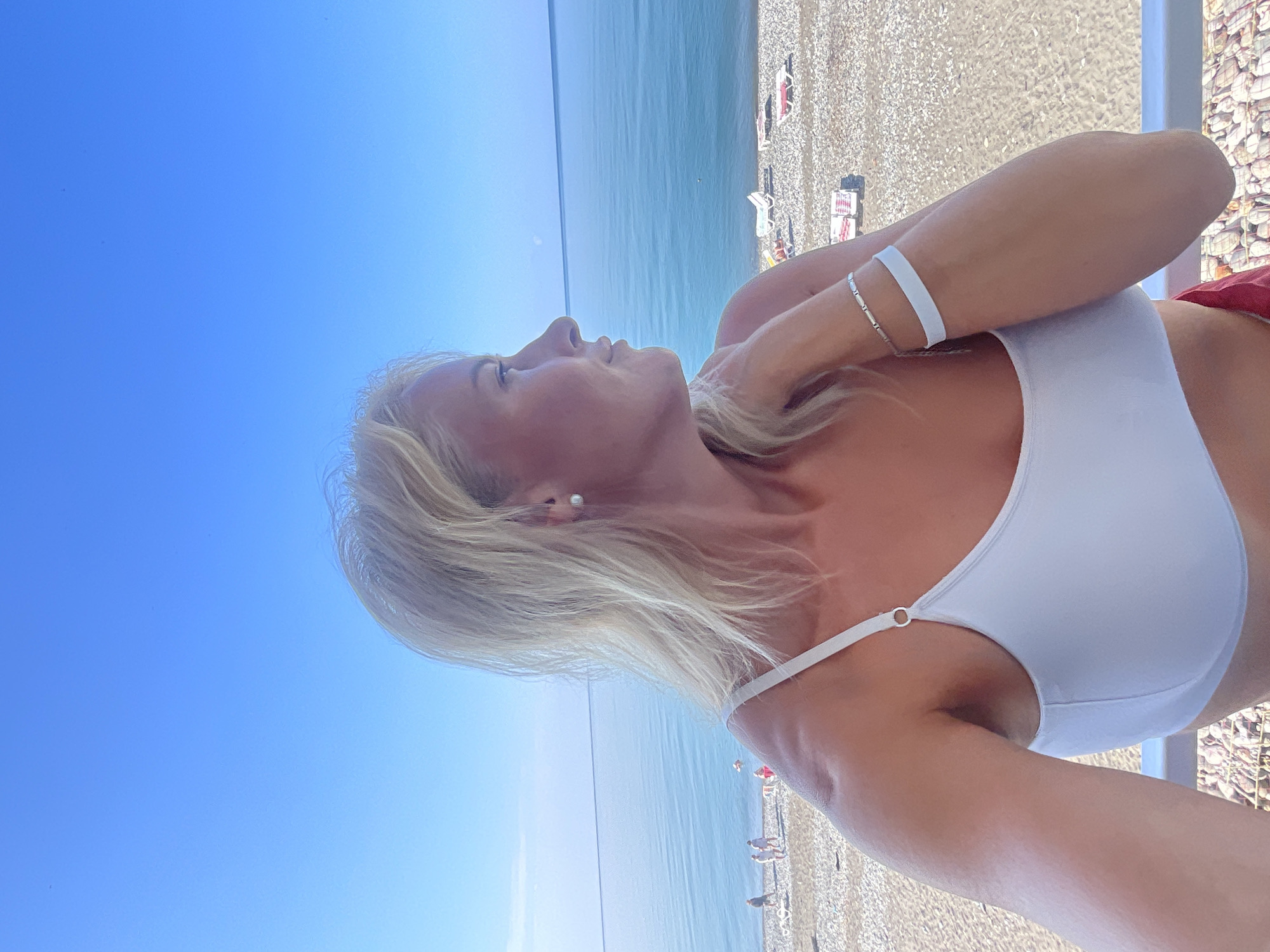 An outdoor, waist-up shot shows a blonde woman with fair skin wearing a white bra top on a balcony overlooking a pebble beach and the sea. The woman is positioned in the right half of the frame, facing left in profile with her hair down. She has on a pearl earring and a silver bracelet and a white band on her left wrist. In the background, the sea is calm and a clear blue sky stretches above. The beach has a few people walking and lounge chairs scattered on the pebbles. The location is Giachrypsh, Abkhazia.