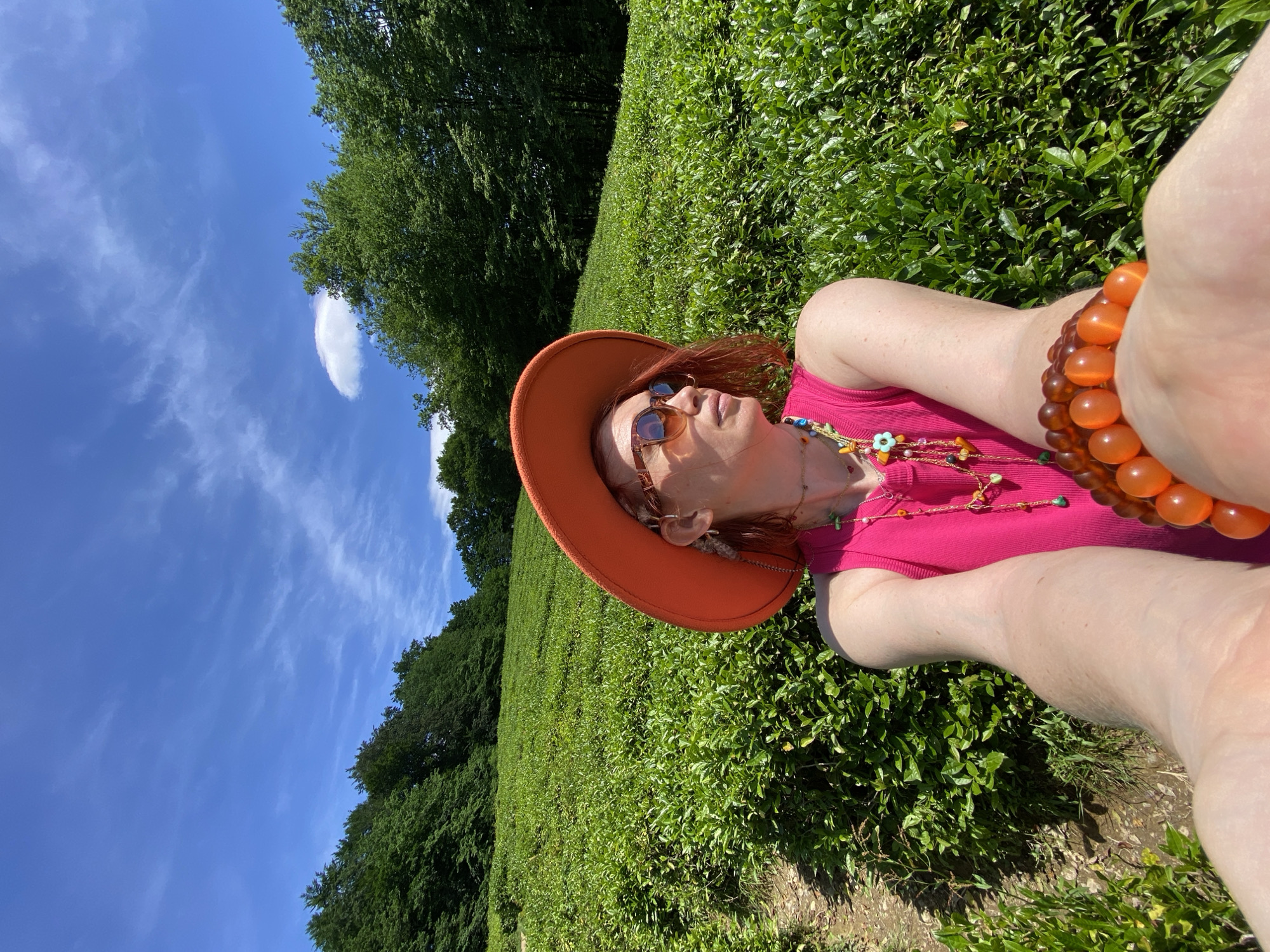 A woman in a pink top and orange hat is lying in a green field. She is wearing sunglasses and orange bracelets. The field is adjacent to a forest, under a blue sky with sparse clouds. The exact location within Izmaylovka, Russia, cannot be determined from the image.