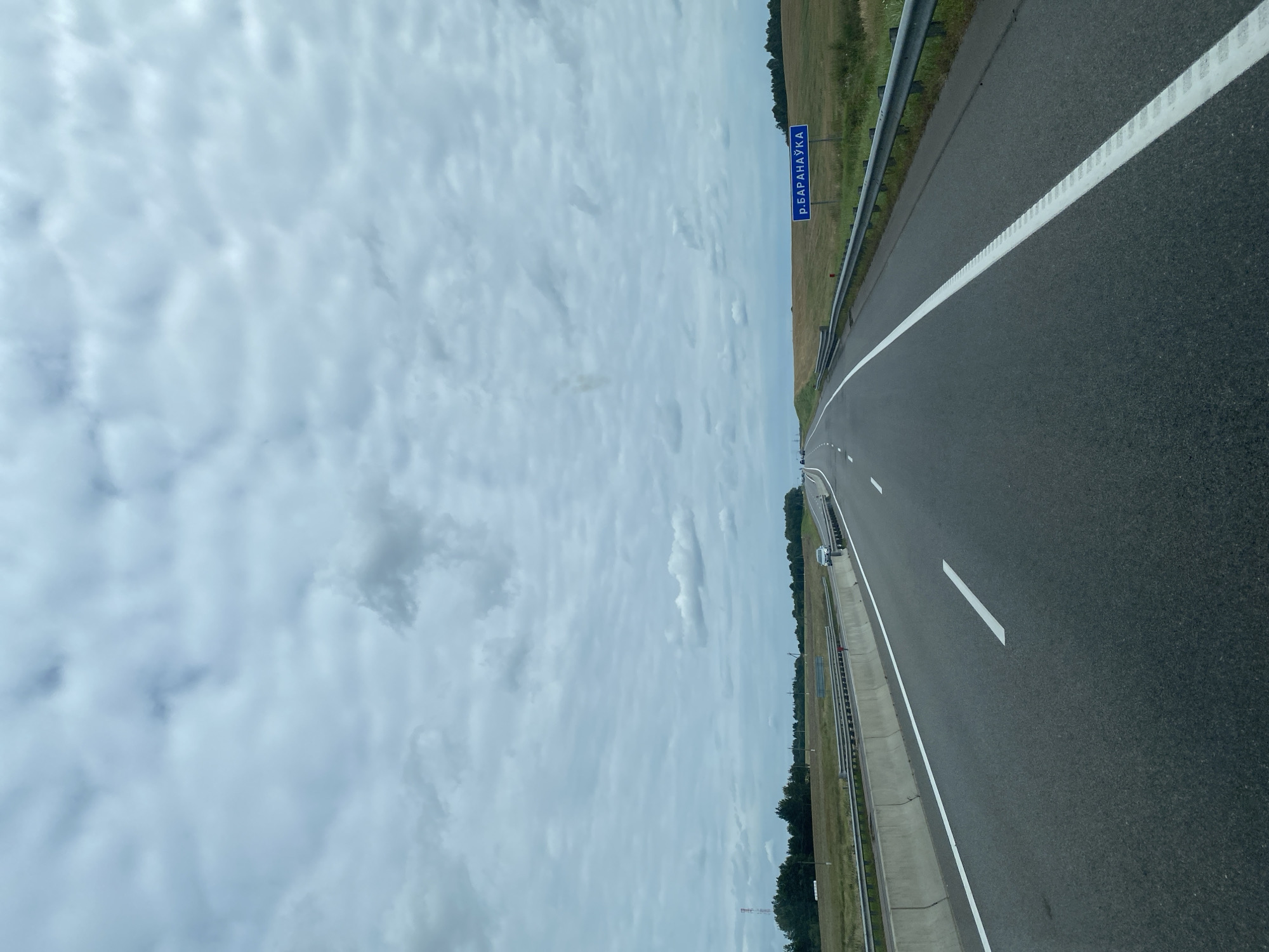 A forward-facing, eye-level shot captures an asphalt highway under a cloudy sky, likely in the Valozhyn District of Belarus. The road curves gently to the right, with a concrete barrier and a metal guardrail lining the left side. White lane markings are visible on the dark gray asphalt. Fields of dry grass and distant trees border the highway. A blue sign with white Cyrillic text reading "р. БАРАНАЎКА" (R. Baranaŭka) is visible on the right side of the road, indicating a location. Several cars are present on the highway in the distance, and a cell tower stands near the treeline on the left. The overall atmosphere is overcast and somewhat muted.