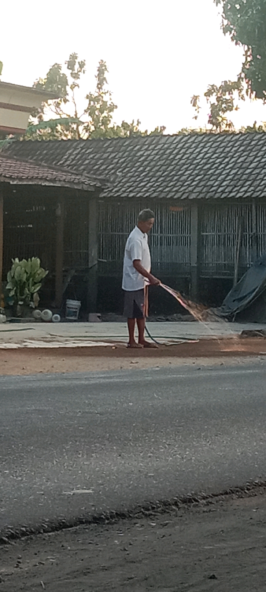 In Kras, Indonesia, a man in a white short-sleeved shirt and gray shorts is watering the ground with a hose. He is standing on a dirt patch next to a paved road. Behind him, a building with a tiled roof and wooden structure is visible. There are trees and foliage in the background. The scene appears to be outdoors during daylight.